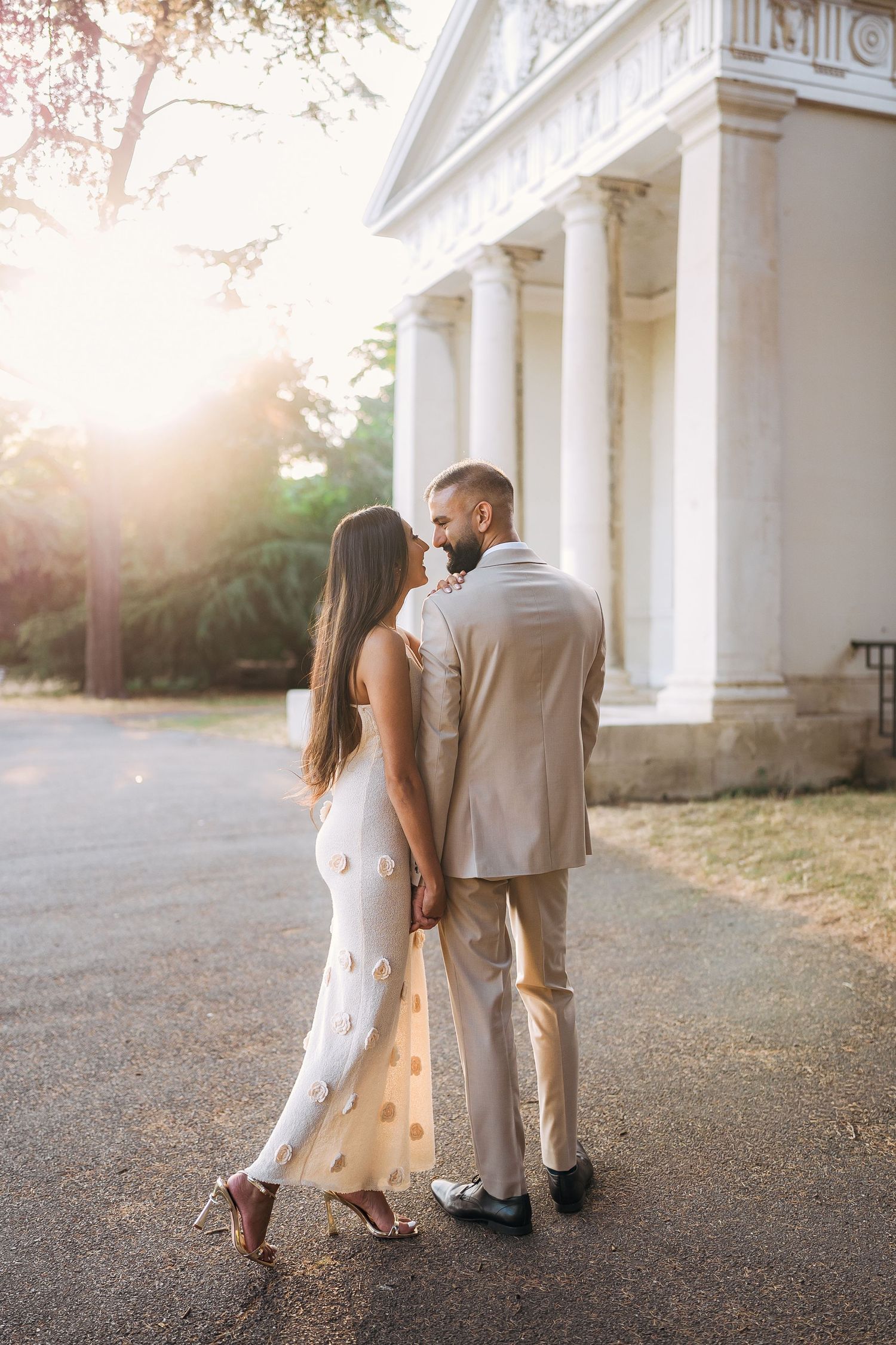 Couple in elegant attire holding hands near a sunlit classical building with white columns at golden hour.