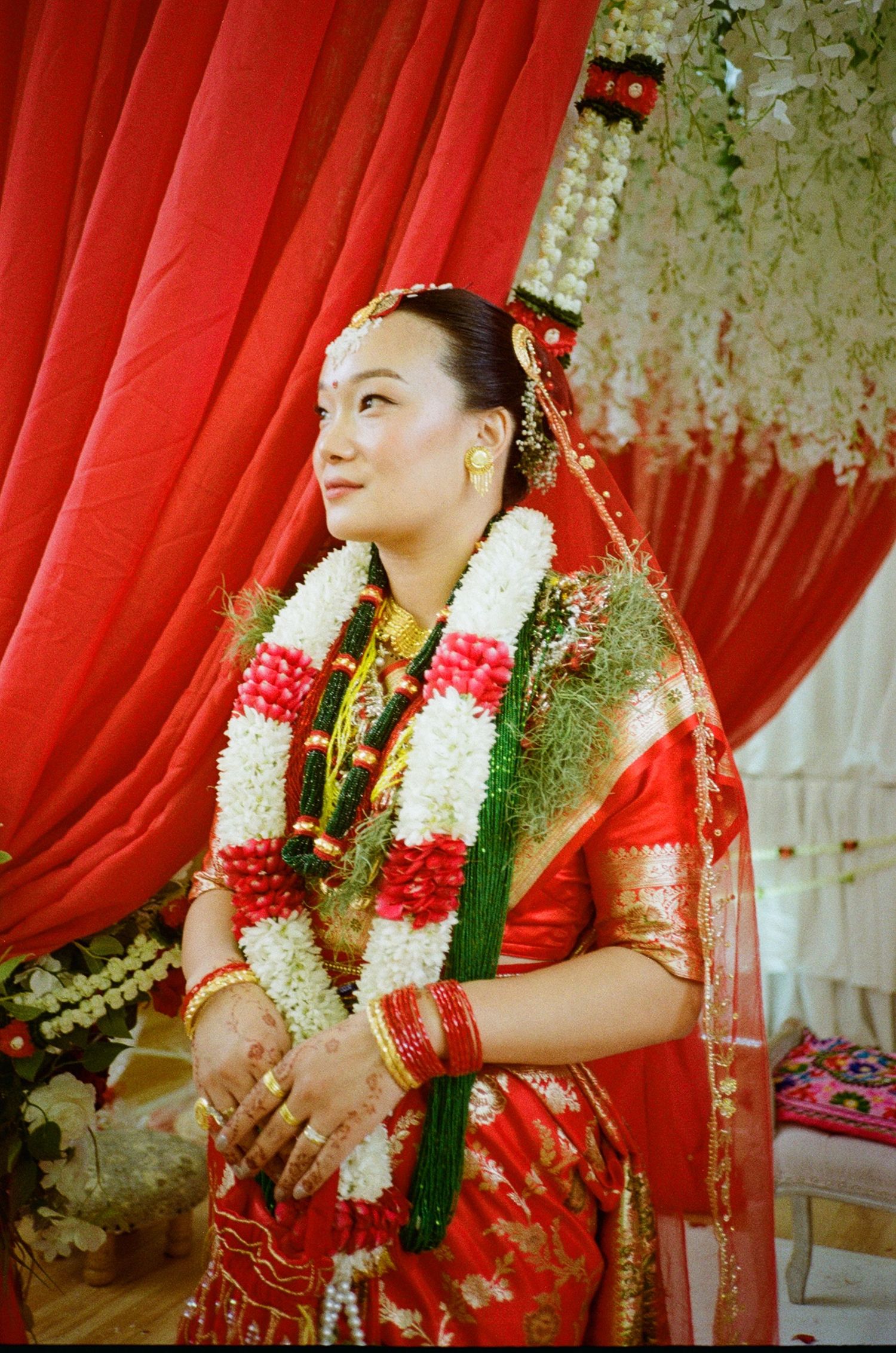 A bride in traditional red saree with floral garlands and gold jewelry stands gracefully against red curtain backdrop.