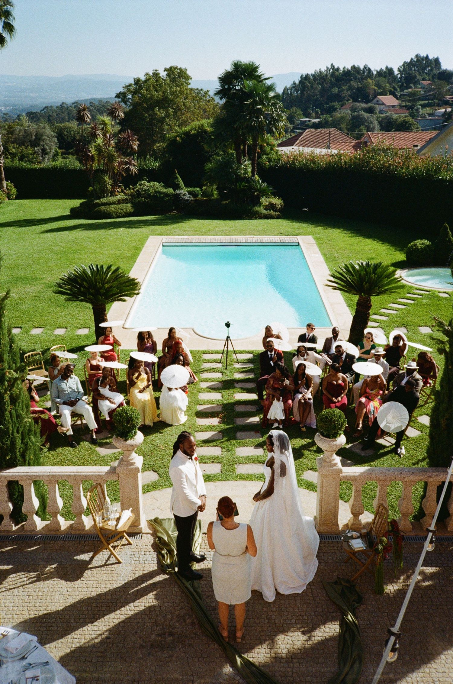 Outdoor wedding ceremony by a pool at a villa, with guests seated holding white parasols and a bride in a white gown.