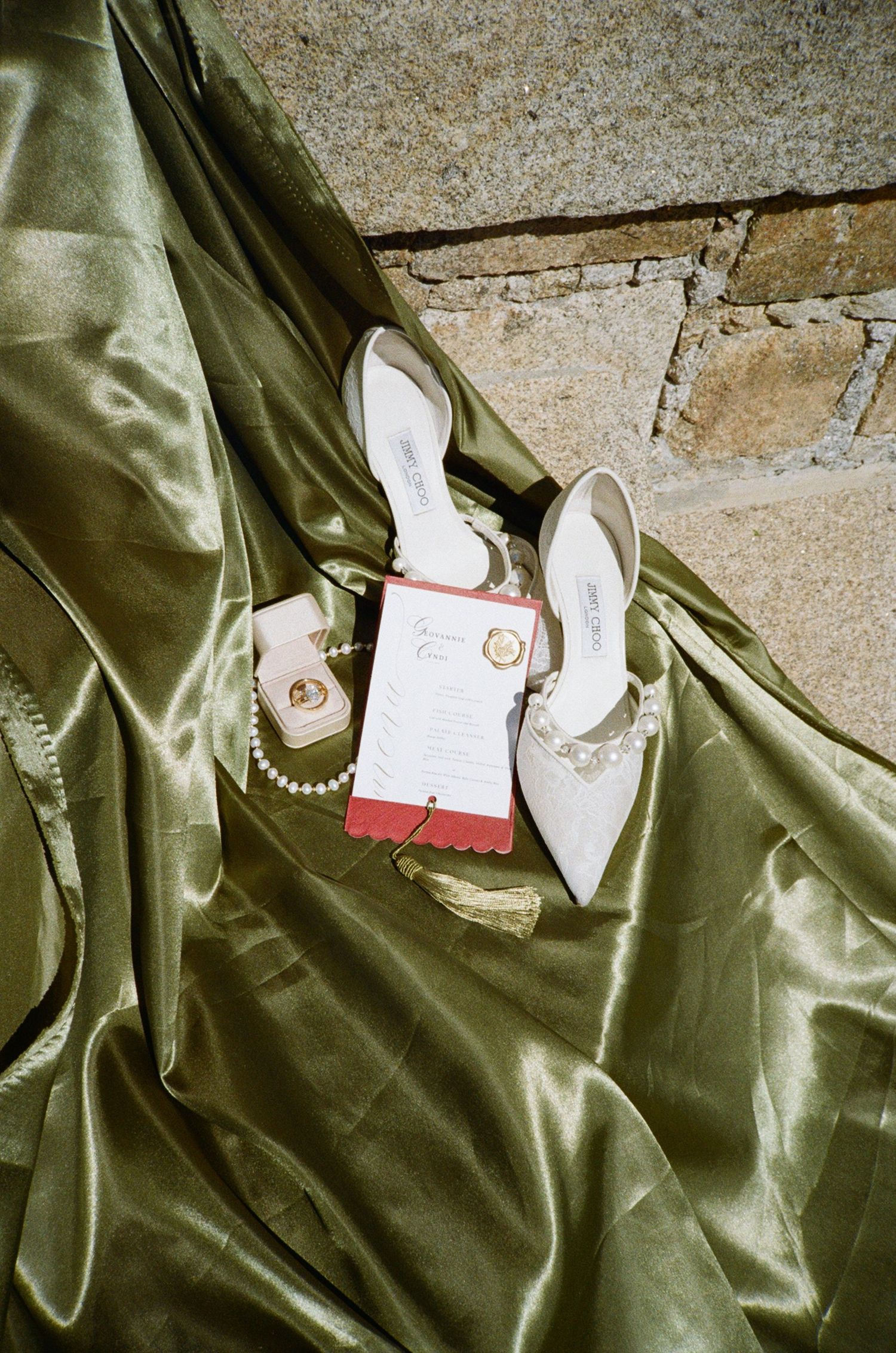 White bridal heels, pearl necklace, gold ring, and red wedding invitation arranged on green satin fabric.
