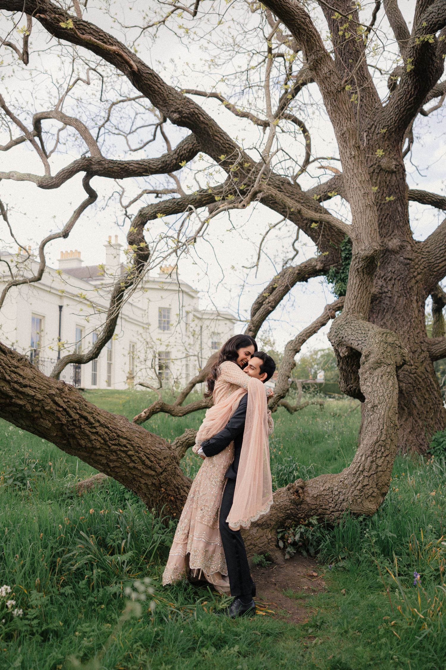 Couple embracing romantically under a large tree in a lush garden with a white manor house in the background.