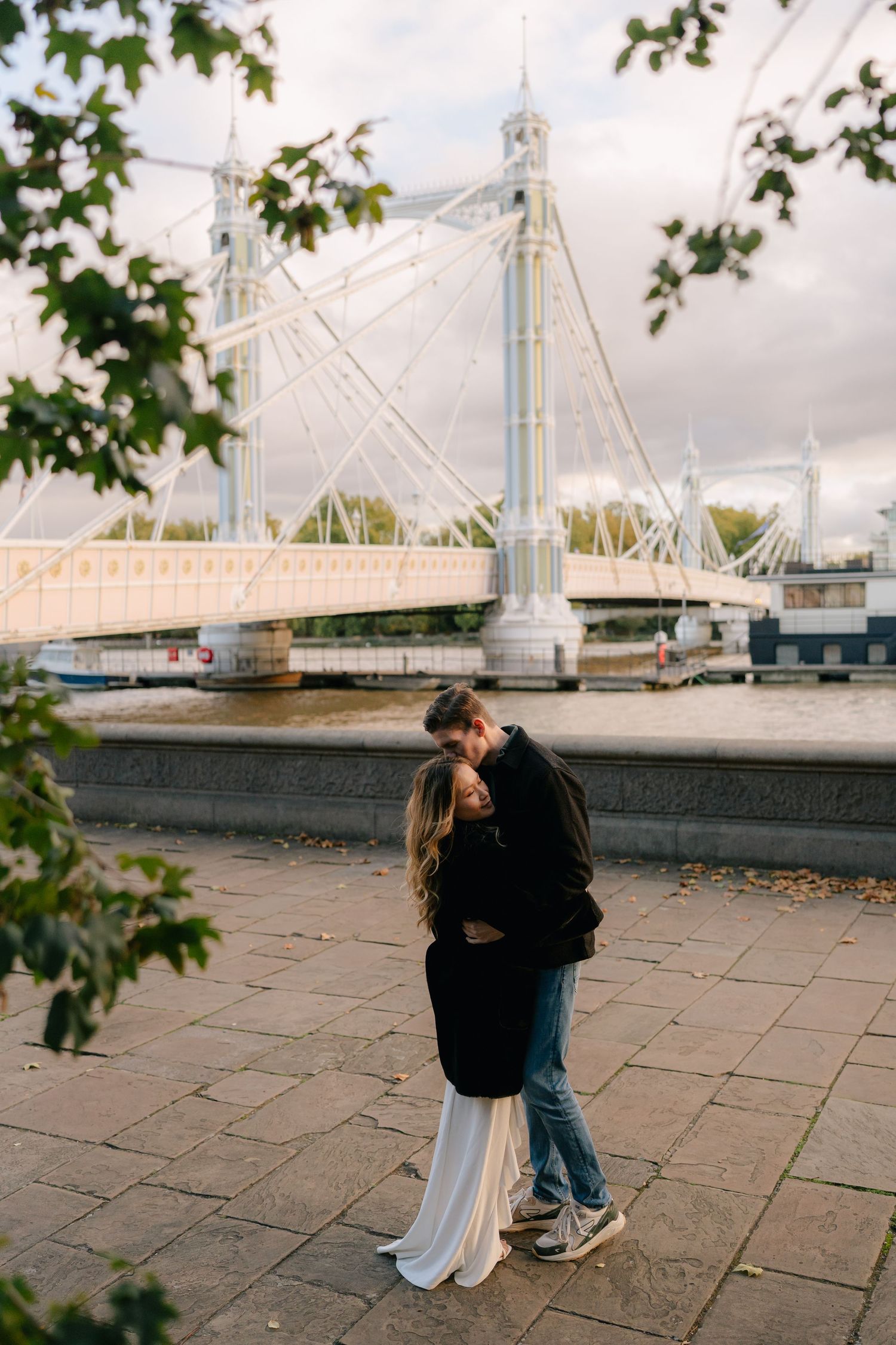 Couple embracing on riverside walkway with white bridge in background.