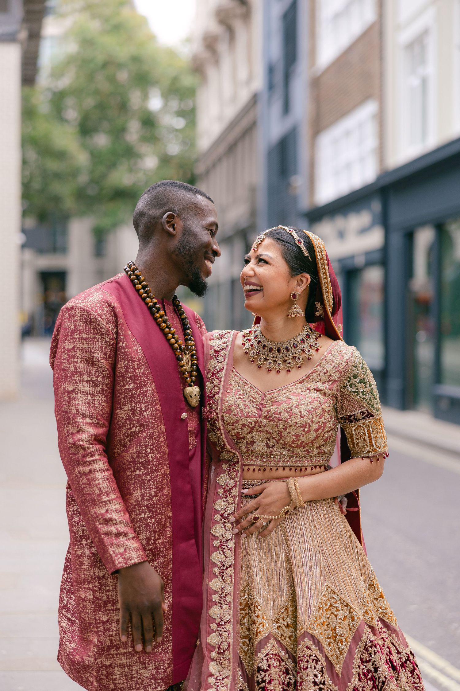 A couple in traditional Indian wedding outfits laughs together on a city street.