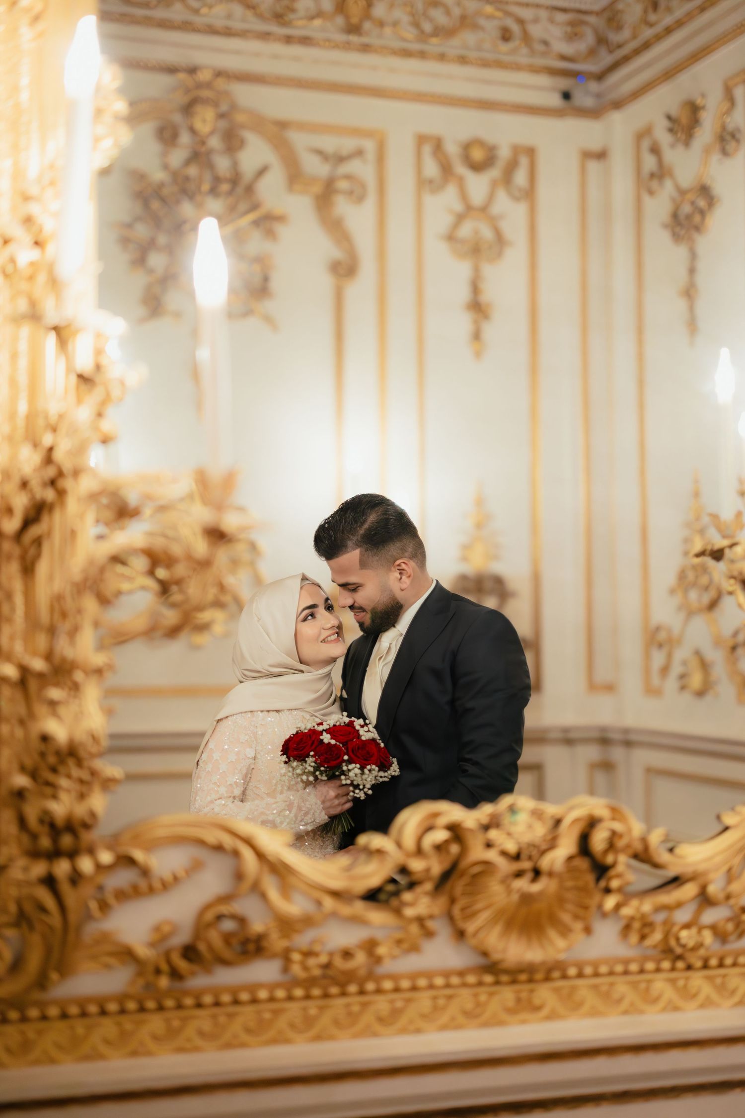 Muslim bride in hijab and groom embrace in ornate gold baroque wedding hall holding red rose bouquet.