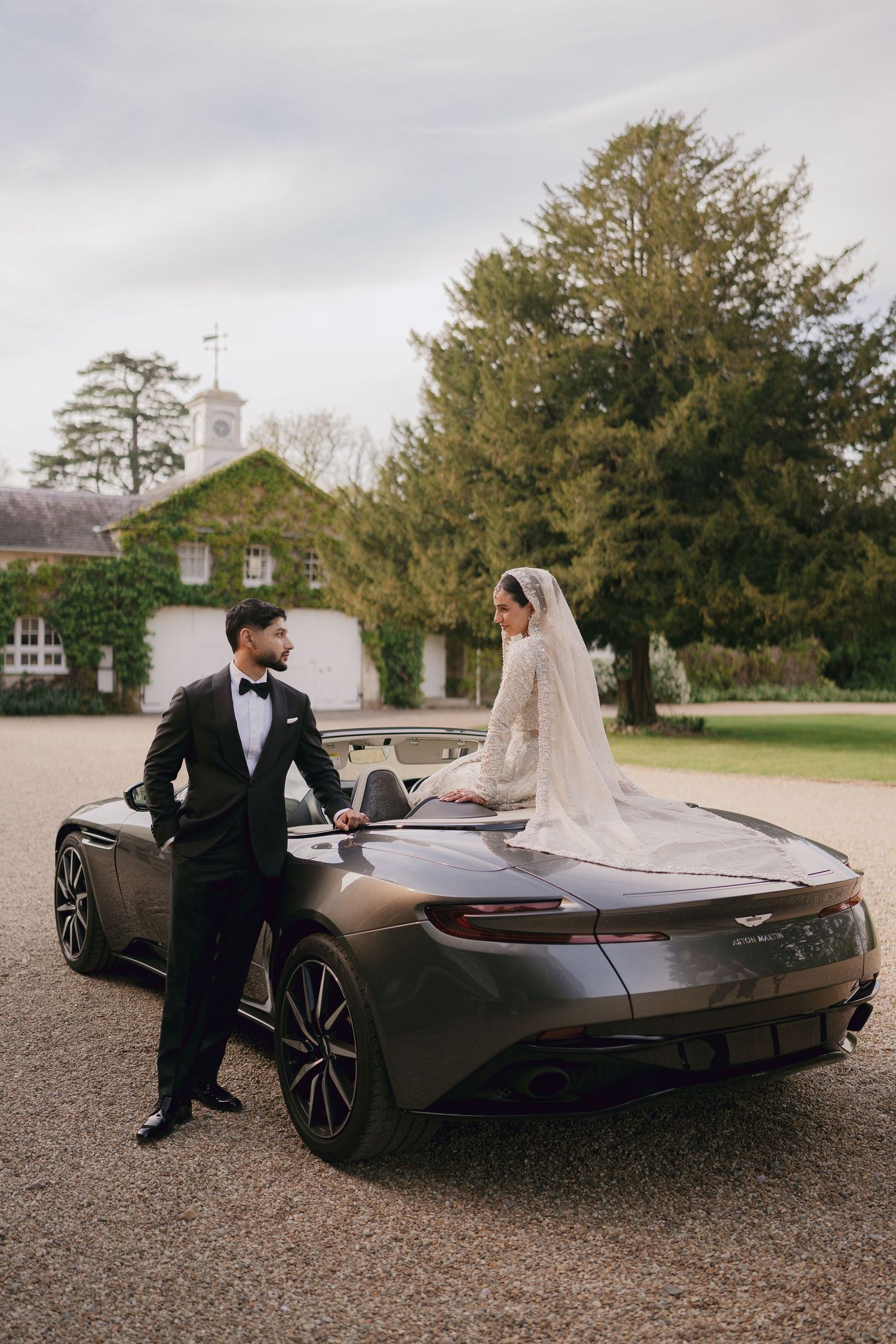 Bride and groom posing beside a luxury Aston Martin at an elegant estate on their wedding day.