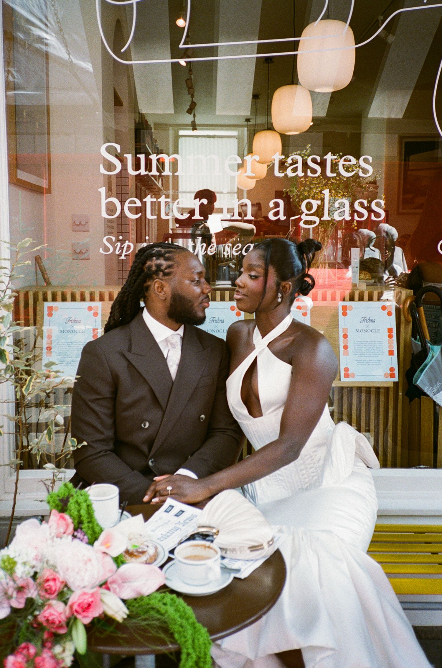 A Black bride and groom sit together at a cafe table, gazing at each other, with pink flowers and coffee cups nearby.