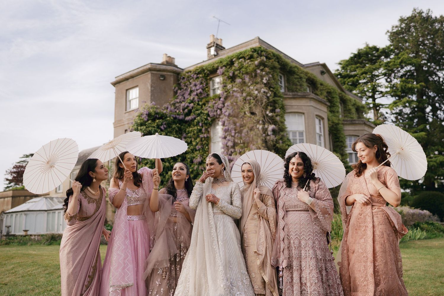 Bride in white lehenga with bridesmaids in pink sarees holding parasols outside ivy-covered English manor.