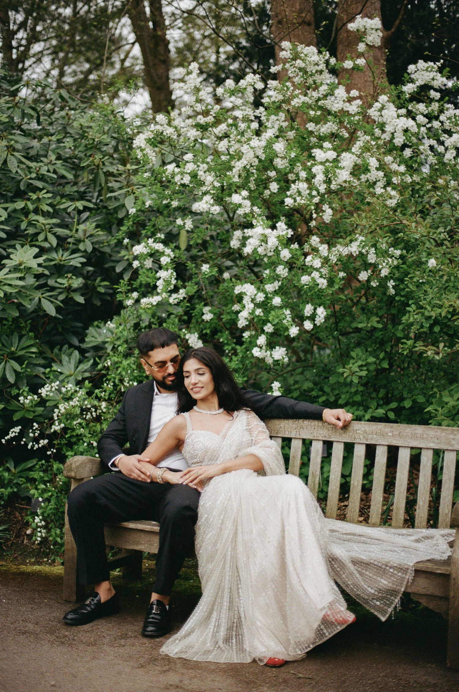 Couple in wedding attire sitting on a garden bench surrounded by lush greenery and white blooming flowers.