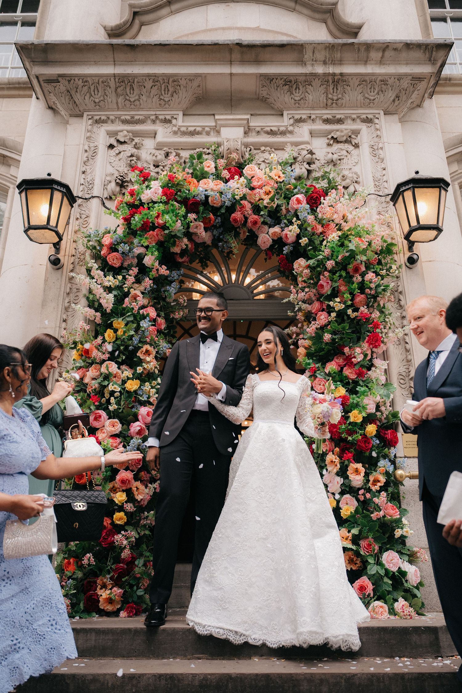 Wedding party sequence captured under stunning pink and red rose arch at classic stone building entrance.