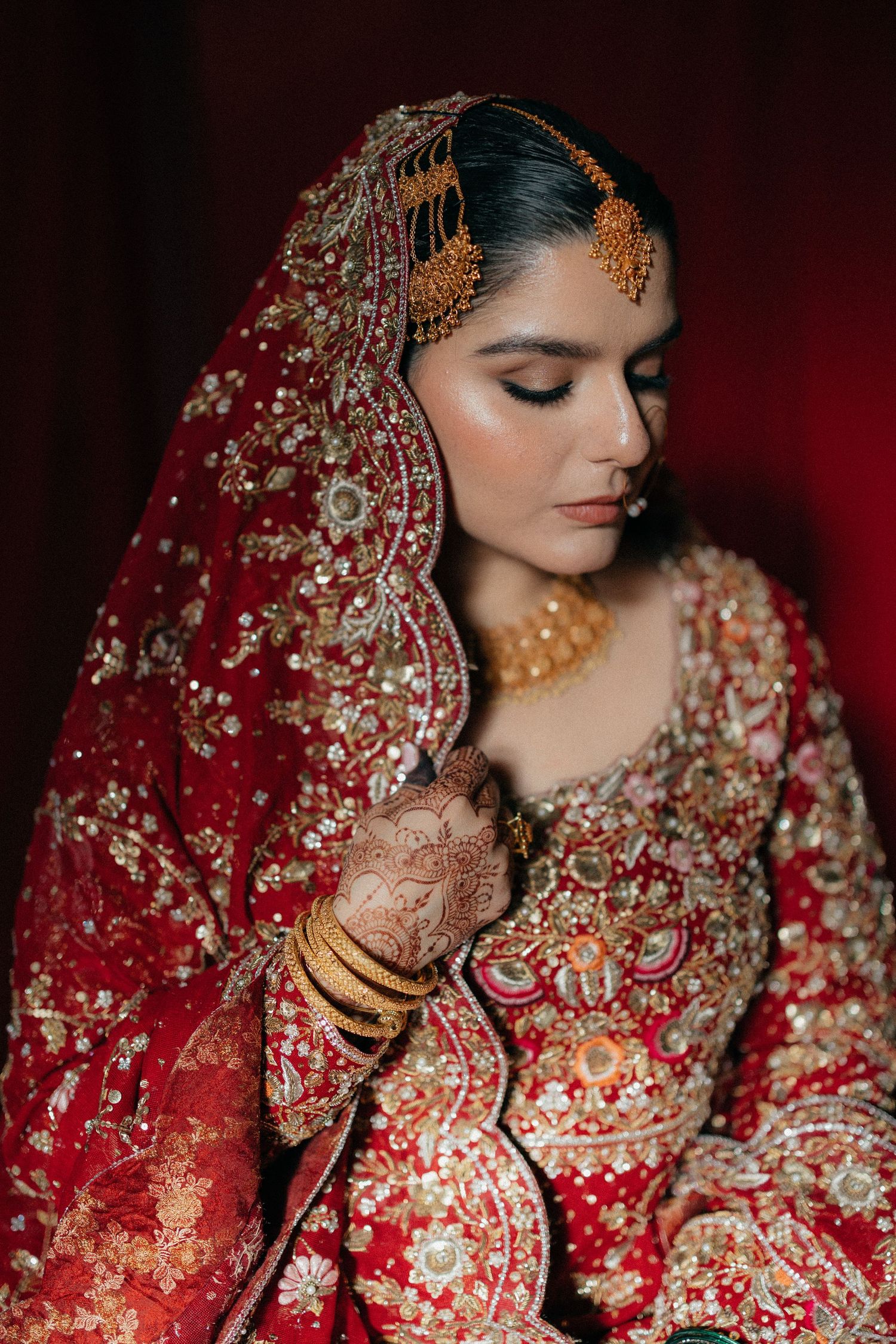 A bride in red embroidered lehenga with gold jewelry, mehndi, and dupatta poses gracefully.