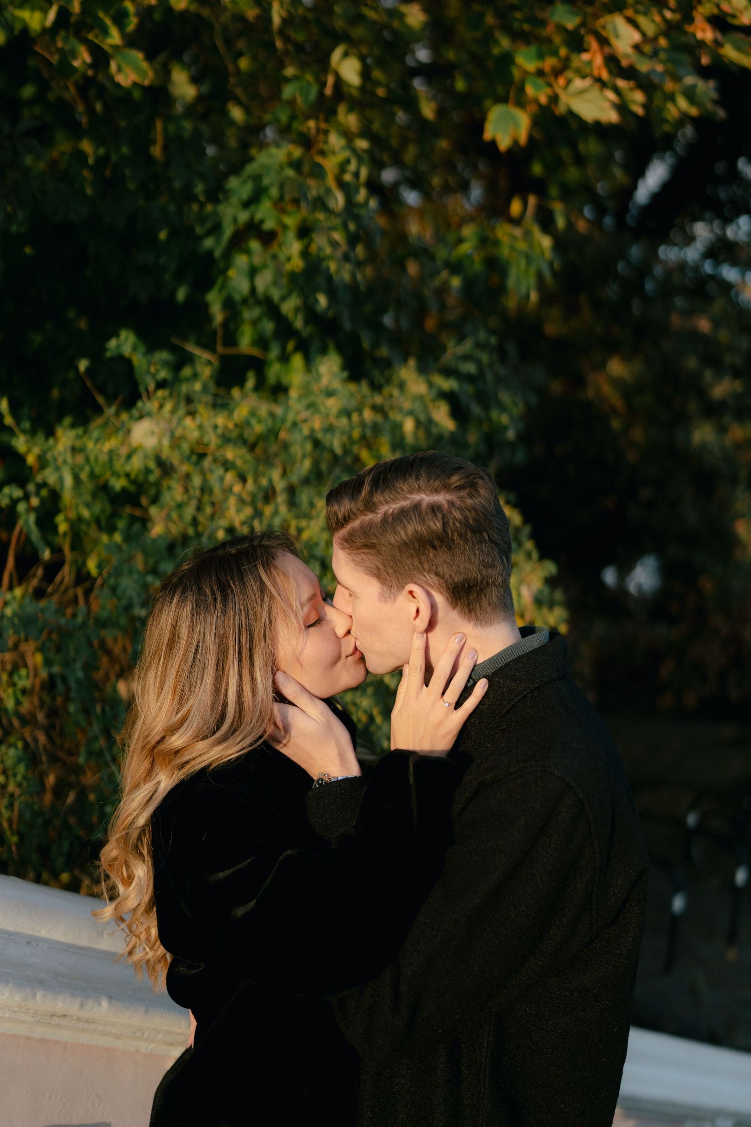 A couple shares a romantic kiss surrounded by autumn foliage in warm sunlight.