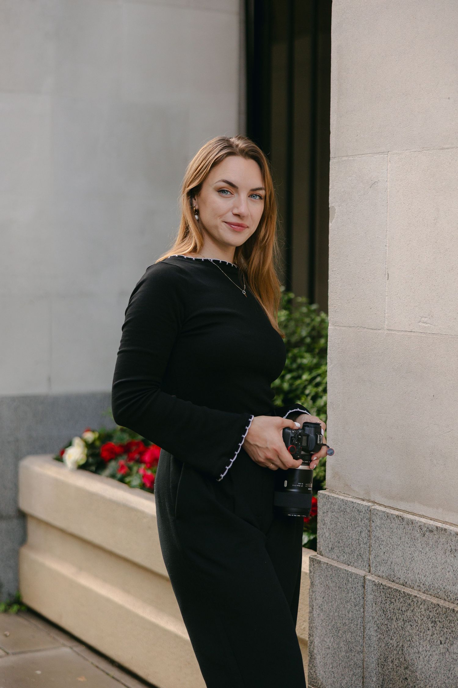 Woman in black velvet dress poses elegantly beside red flowers and stone wall.