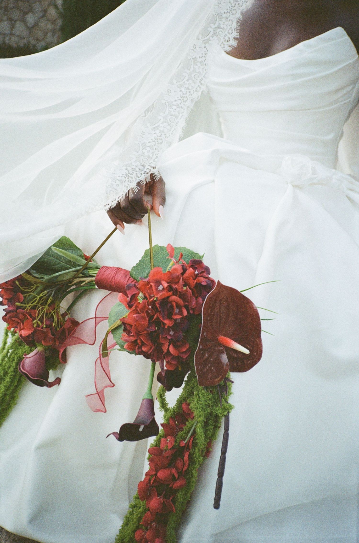 Bride in white wedding gown holding a lush red bouquet with anthuriums, hydrangeas, and moss accents tied with ribbon.