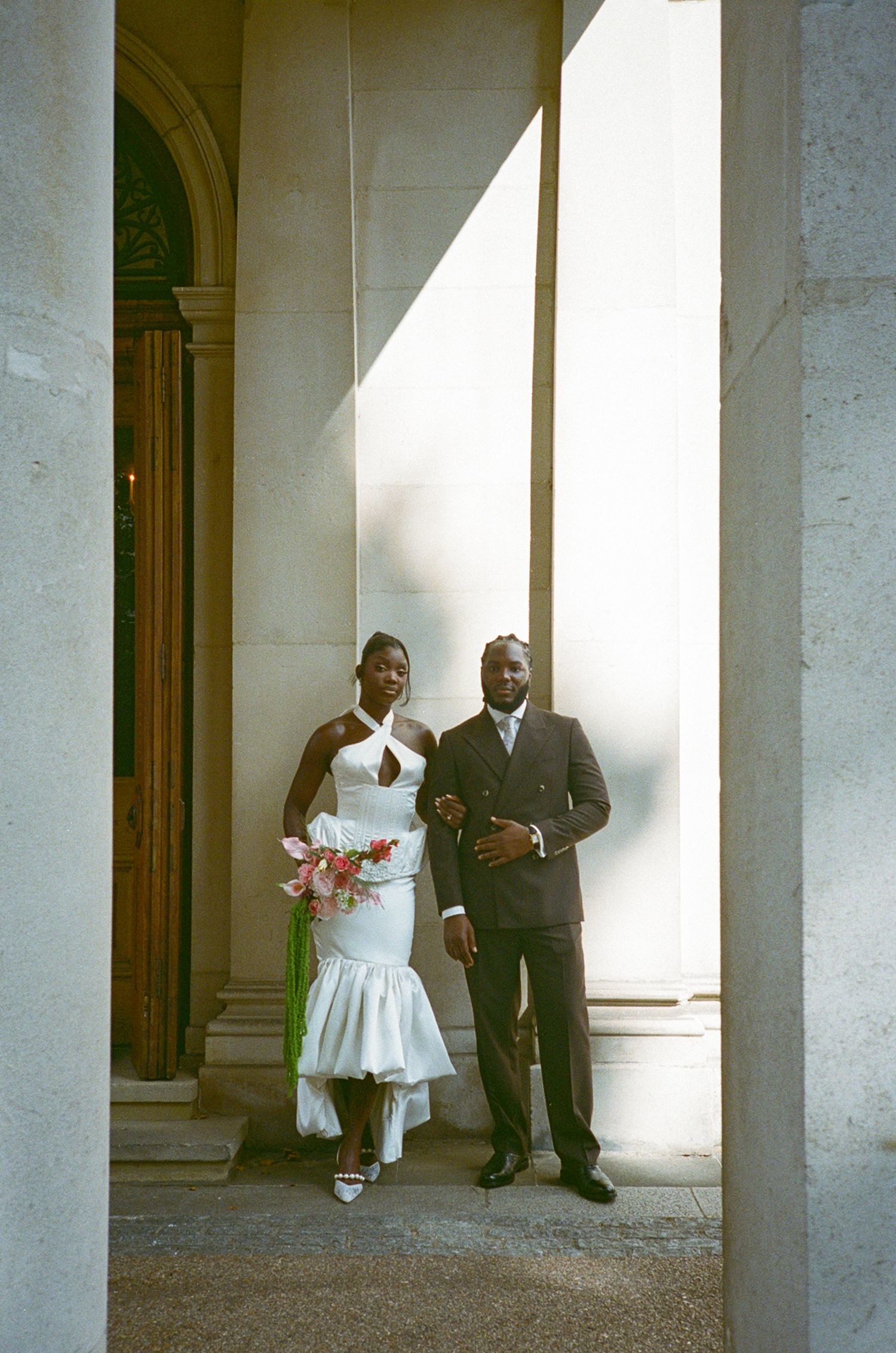 A bride in white halter gown and groom in dark suit pose between columns at an elegant wedding venue.
