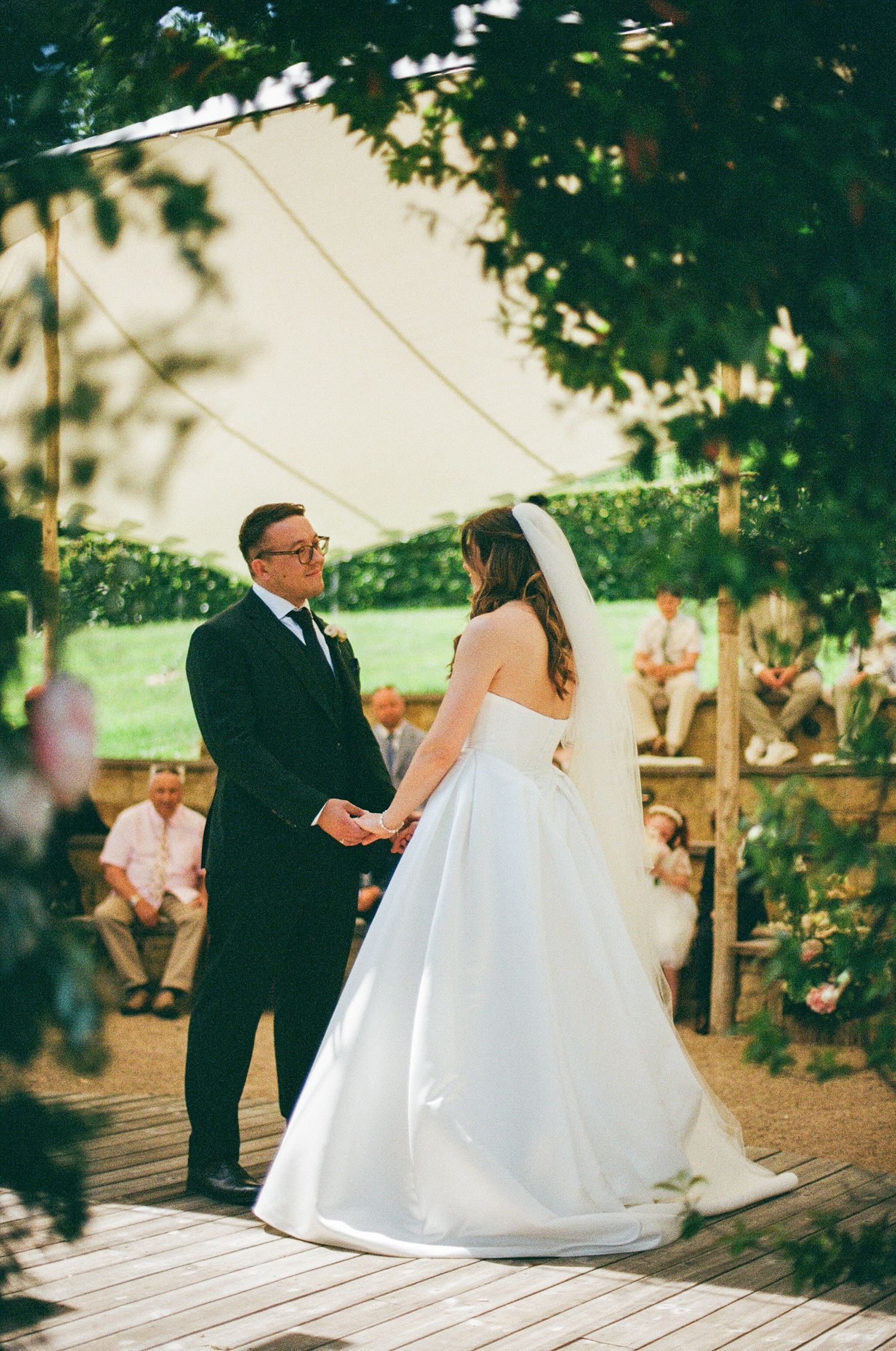 A bride and groom hold hands during an outdoor wedding ceremony under a white tent surrounded by lush greenery.