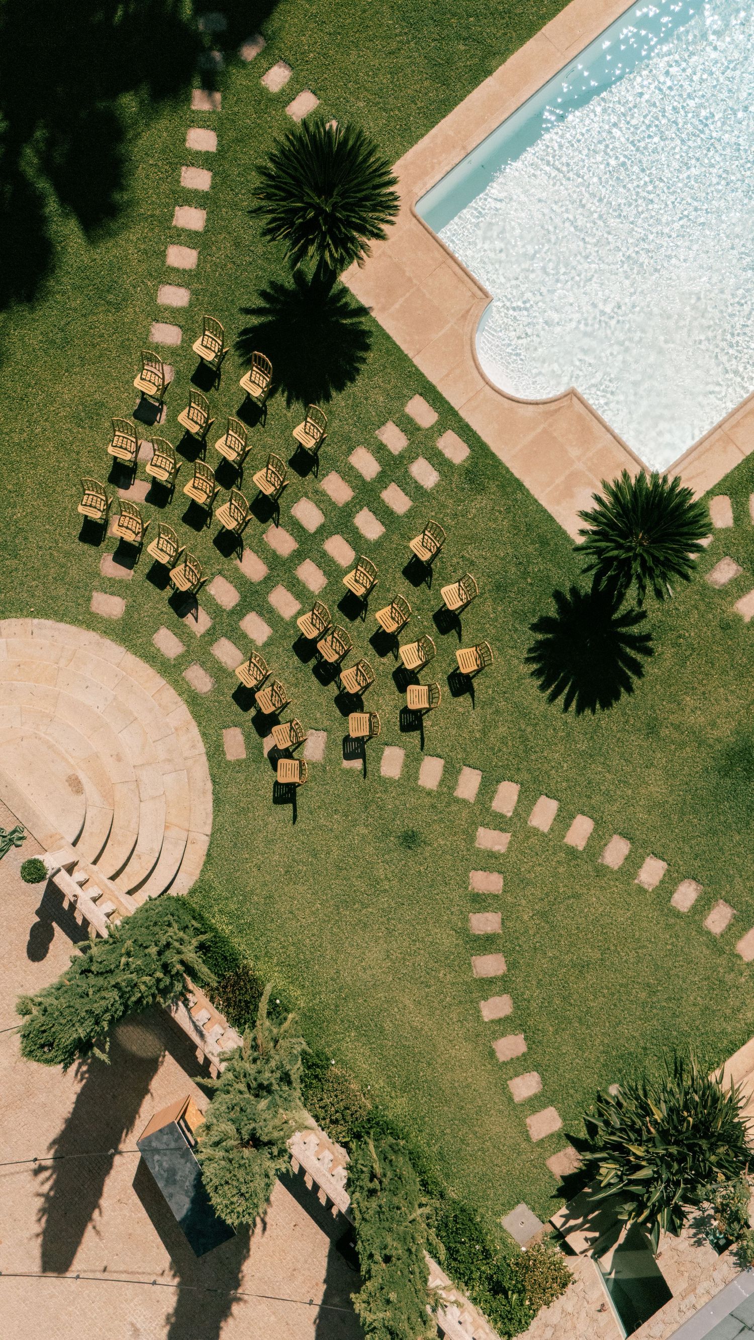 Aerial view of golden chairs arranged on green lawn near a pool, surrounded by palm trees and stone pathways.