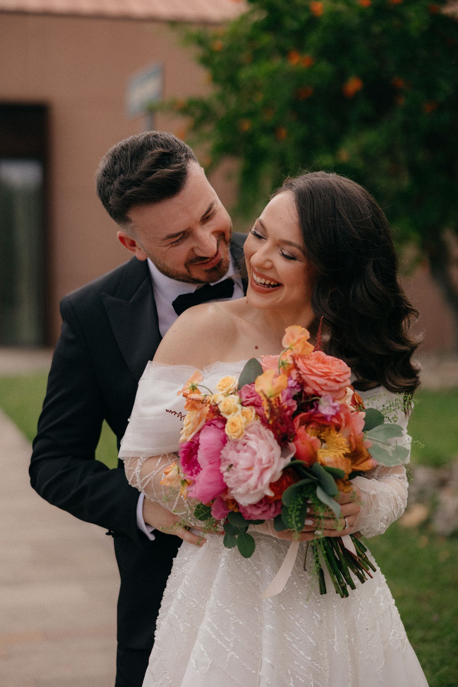 Newlyweds embracing outdoors with vibrant coral and pink floral bouquet.