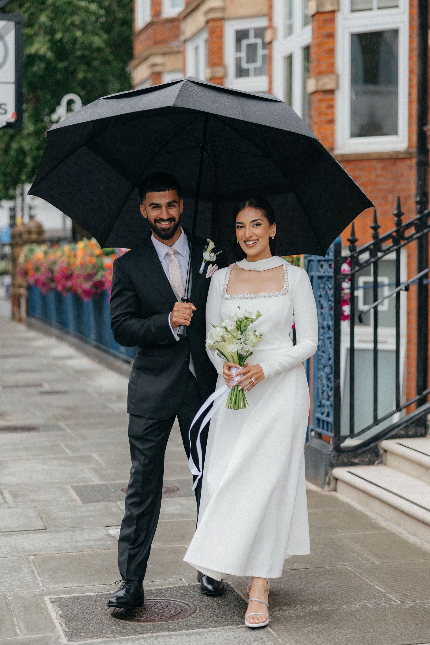 A couple shares black umbrella while walking in formal attire on London street.