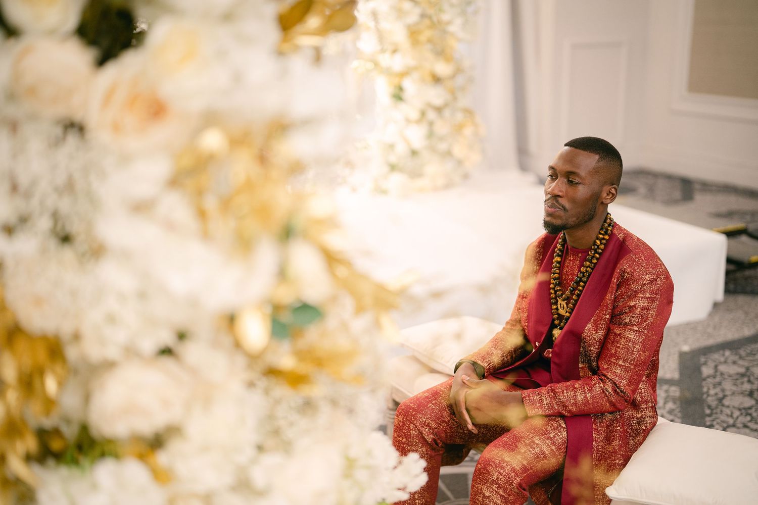 A person in a red traditional outfit sits on steps while smiling and looking off to the side in an outdoor setting.