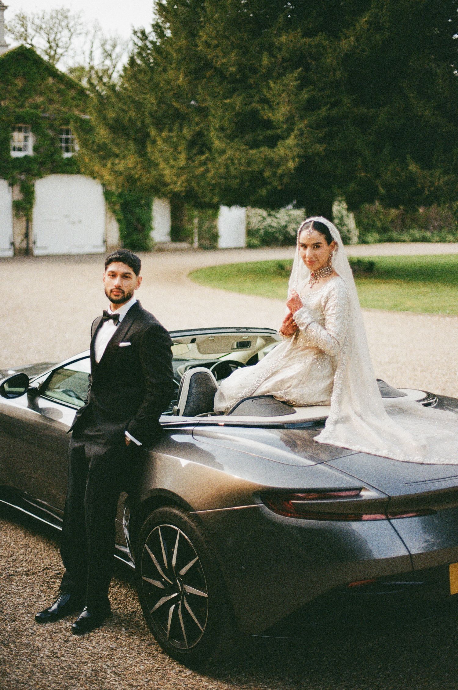 Bride in white lace gown and veil sits atop Aston Martin convertible beside groom in black tuxedo at estate.