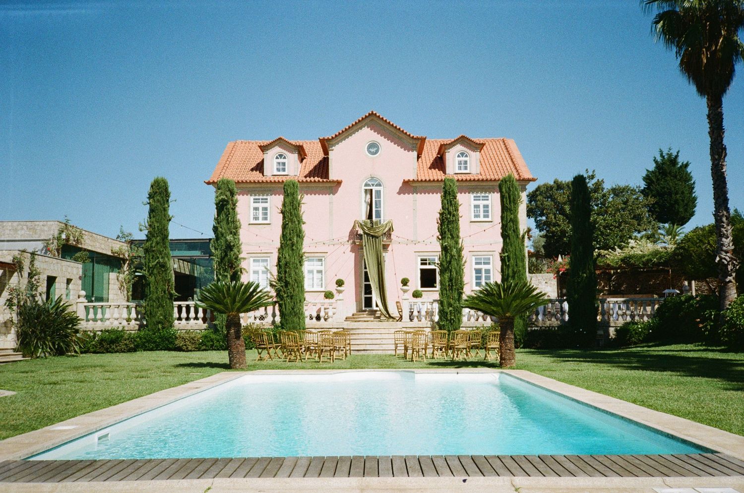 Pink villa with terracotta roof surrounded by lush gardens featuring a large rectangular swimming pool on a sunny day.