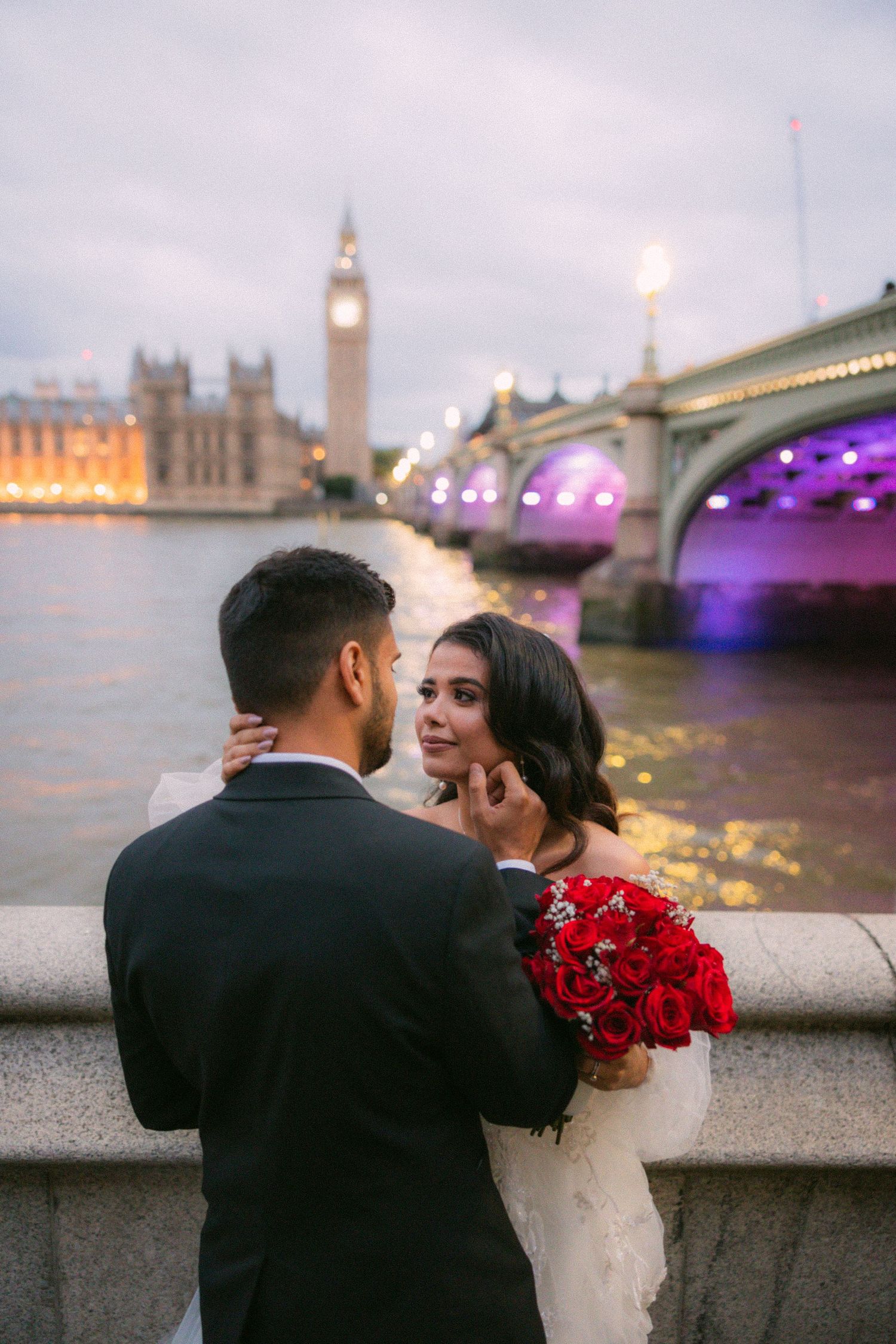 Wedding couple shares intimate moment by Thames River with London landmarks in background at dusk.