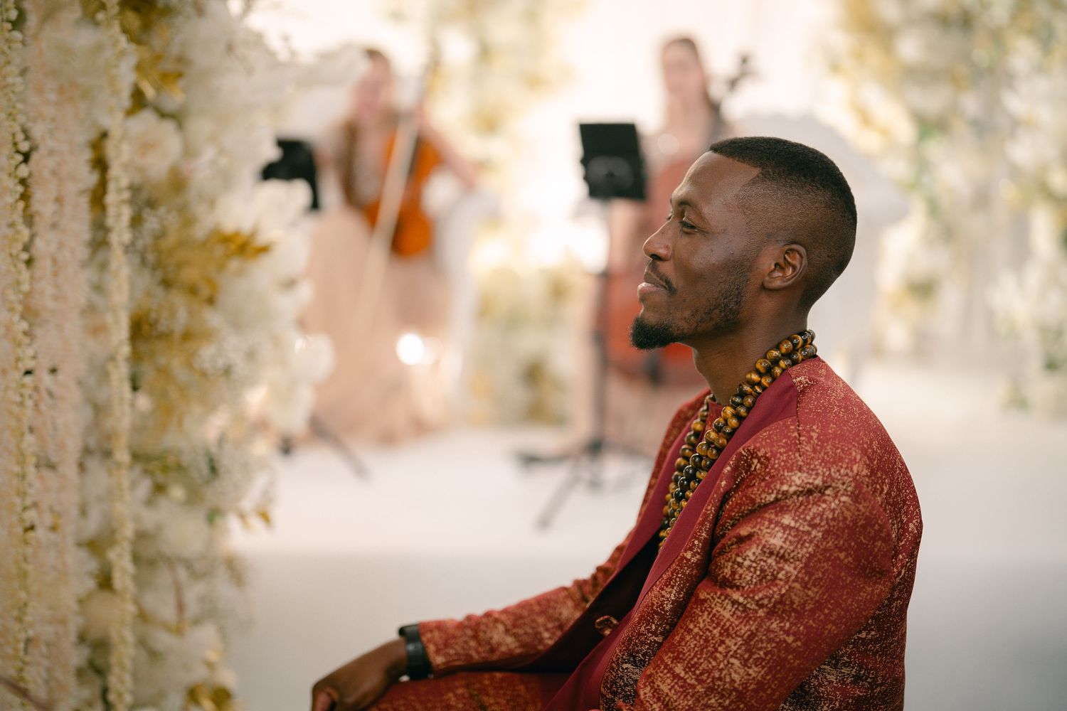 A person in a red and gold traditional outfit sits smiling during a celebratory event with decorative lighting.