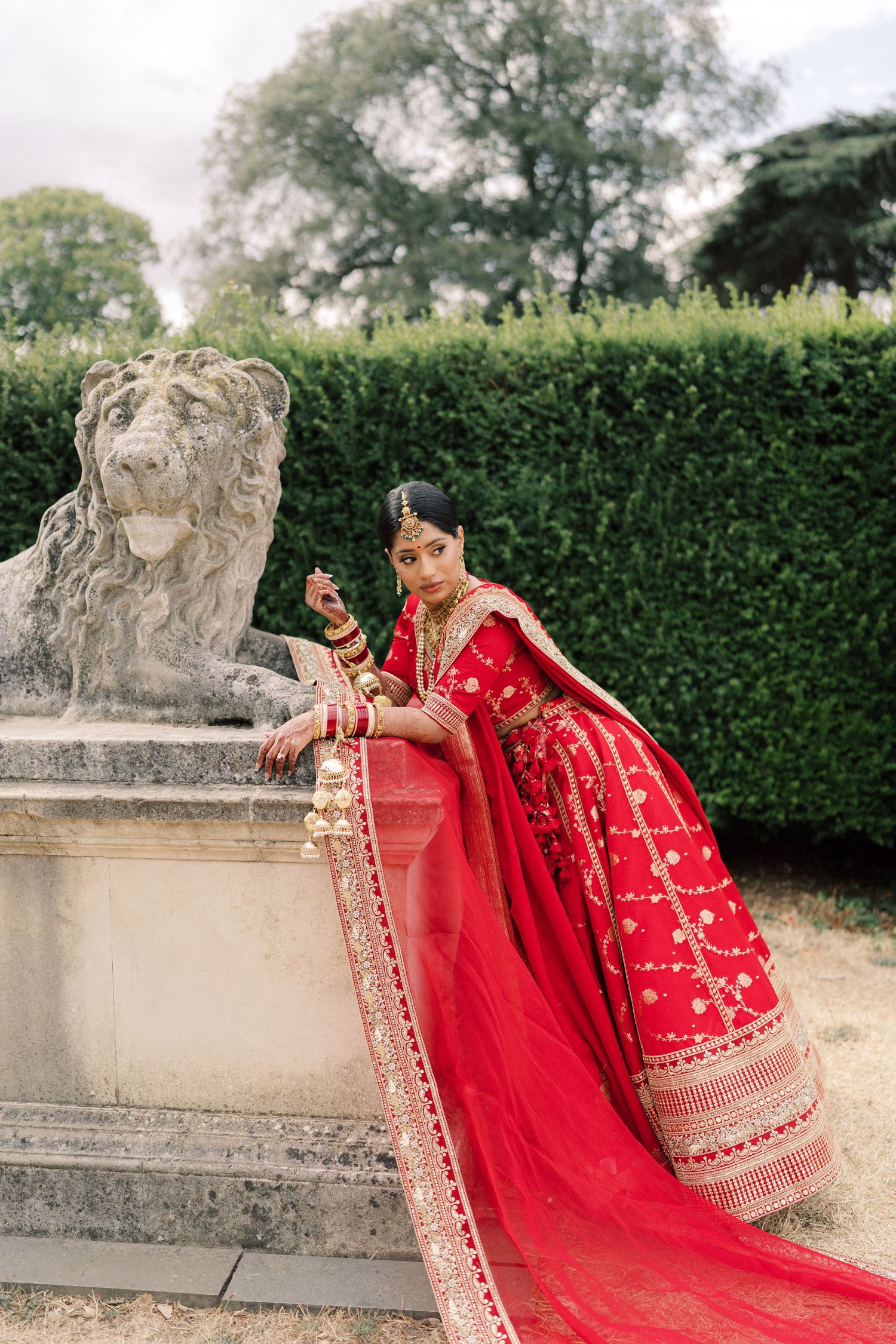 A bride in a red embroidered lehenga poses beside a stone lion sculpture in a lush garden setting.