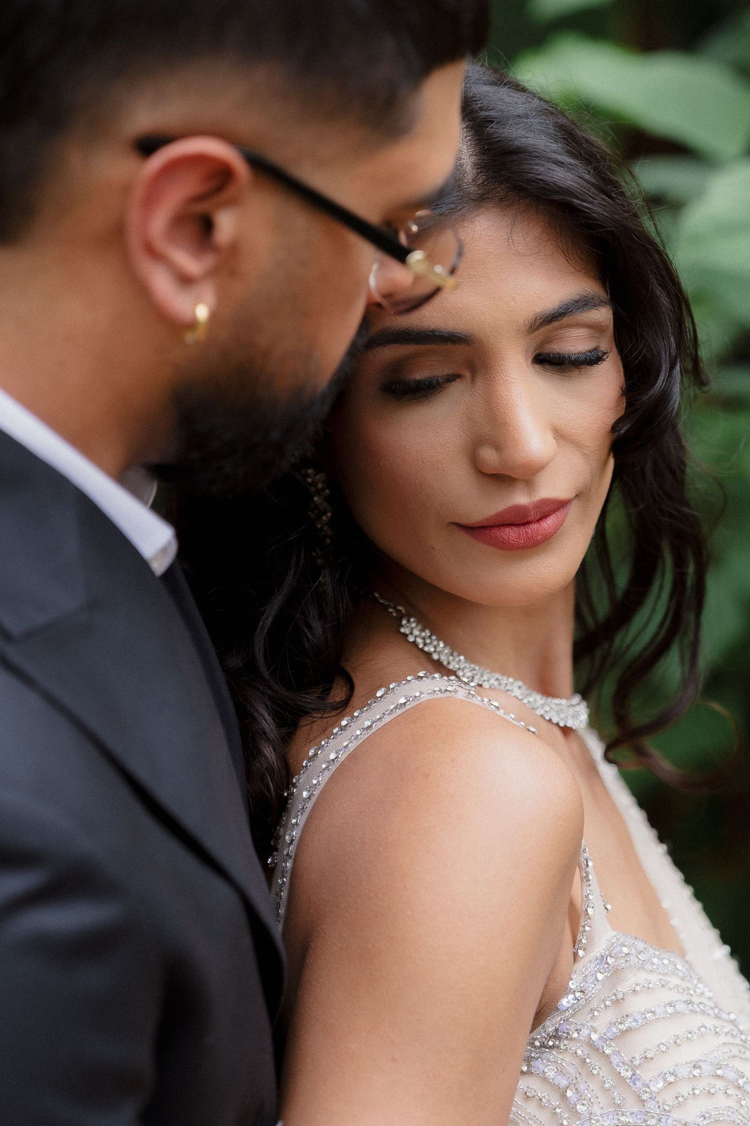 Elegant bride in beaded gown leans against groom in dark suit, sharing an intimate moment outdoors.