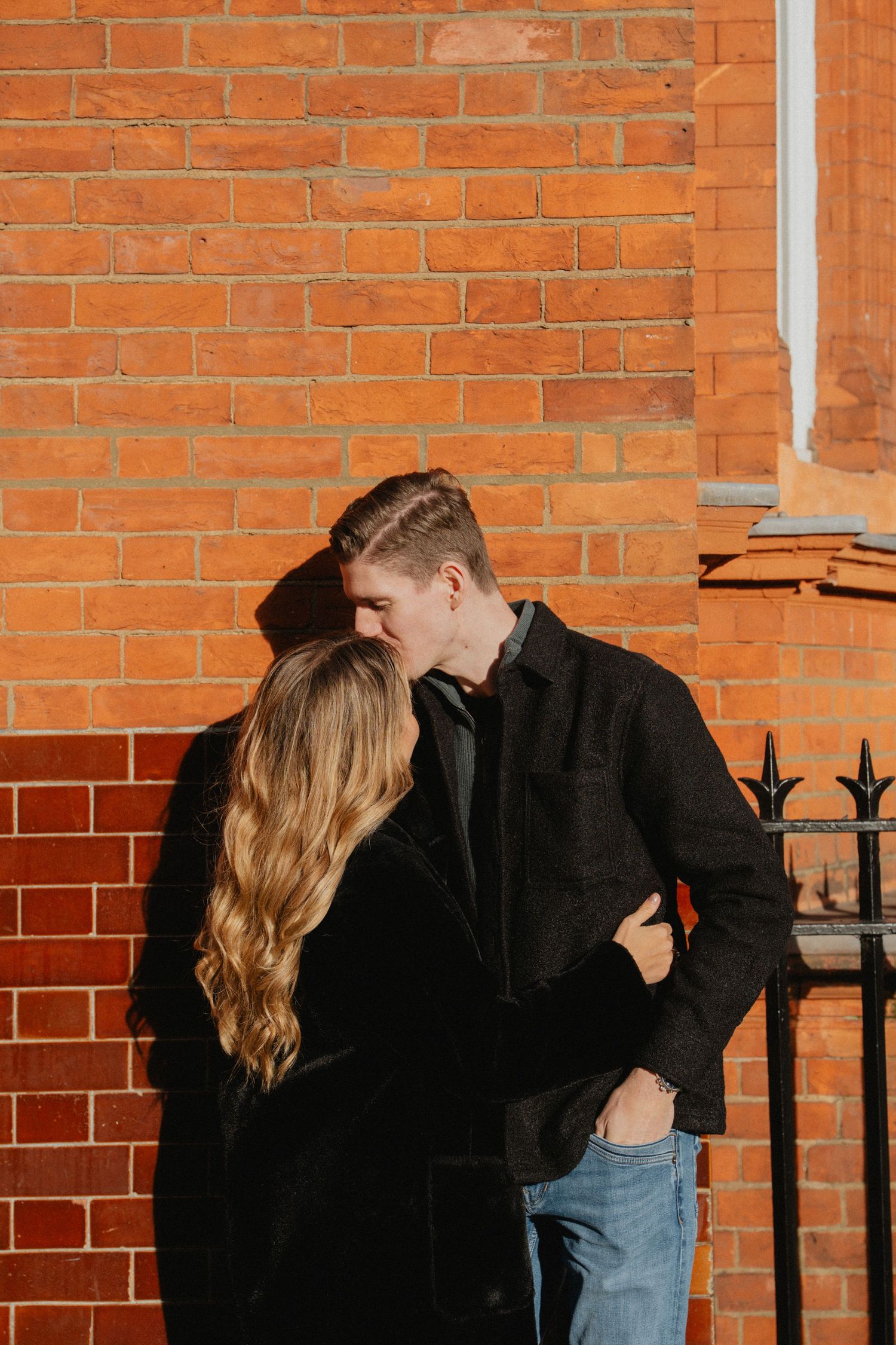Romantic couple embraces against rustic brick wall in urban setting.