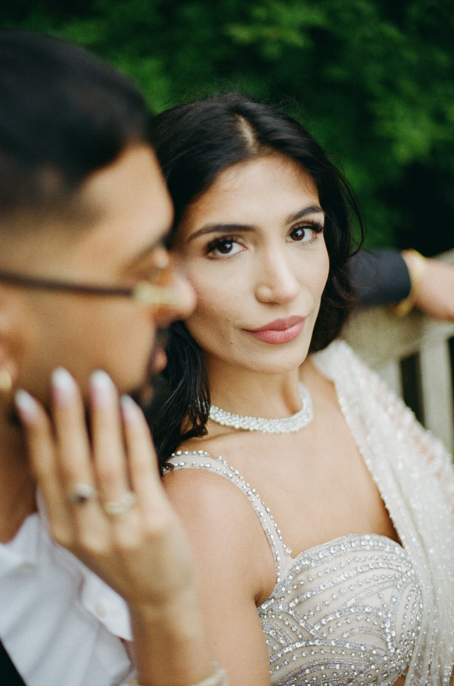 A bride in a beaded gown and crystal choker necklace gazes softly at her partner during a wedding celebration.