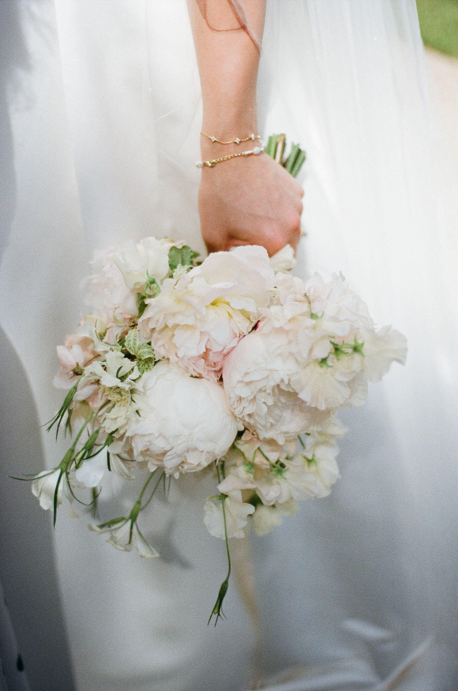 Bride holding a lush white and blush peony wedding bouquet with greenery accents against a white satin gown.