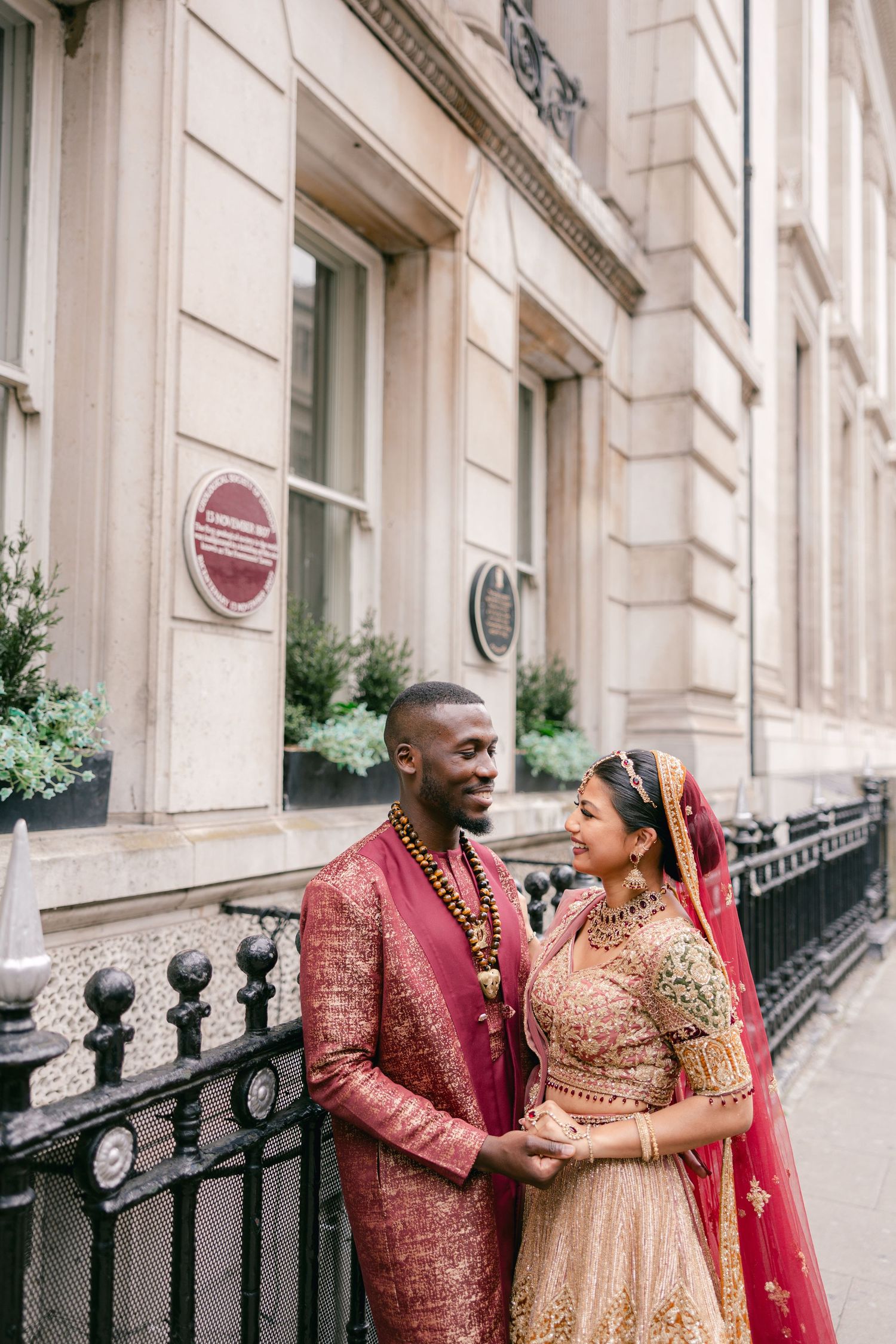 A couple in burgundy and gold Indian wedding outfits laughs together on a classic city street.