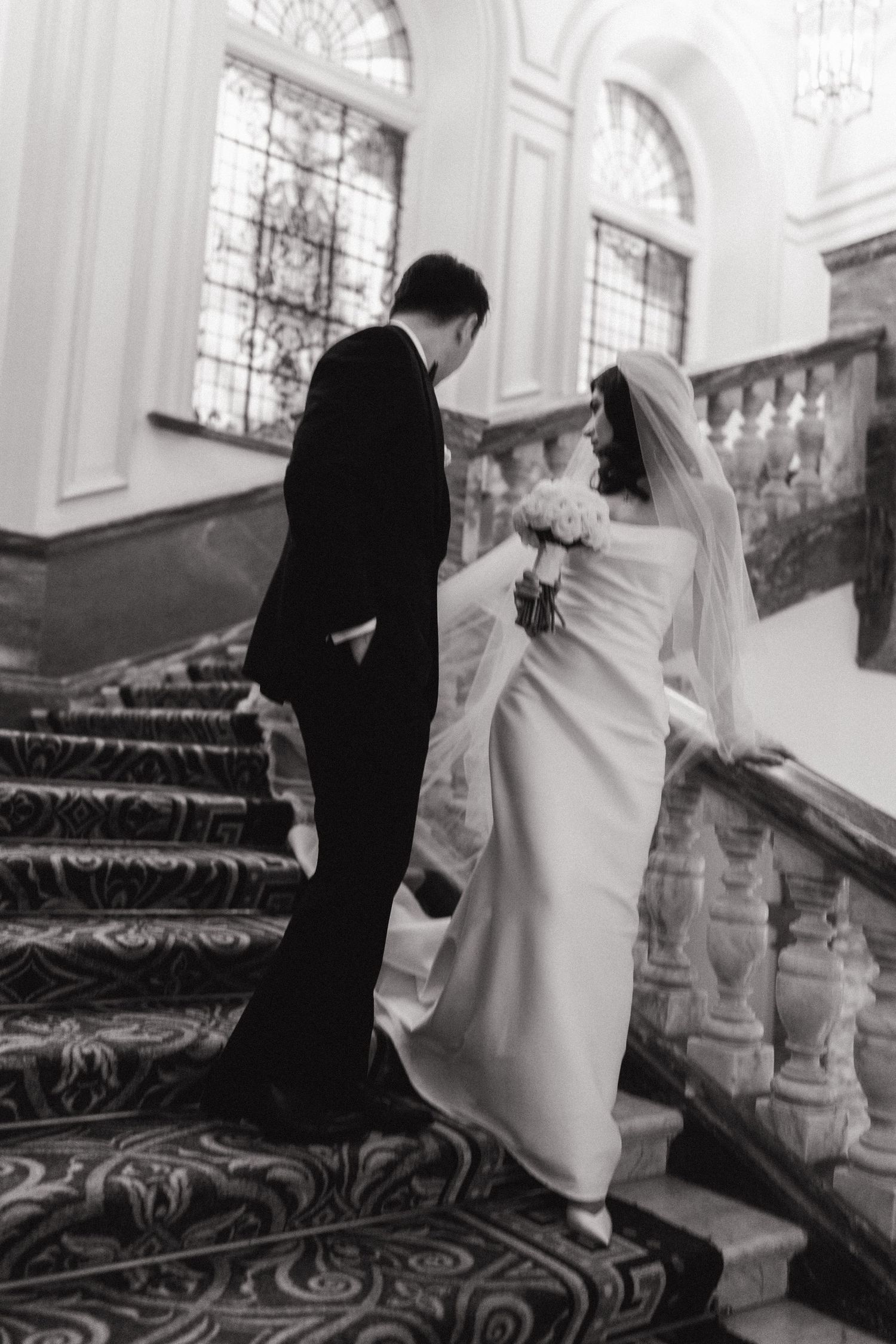Bride and groom ascending ornate staircase with decorative carpet.