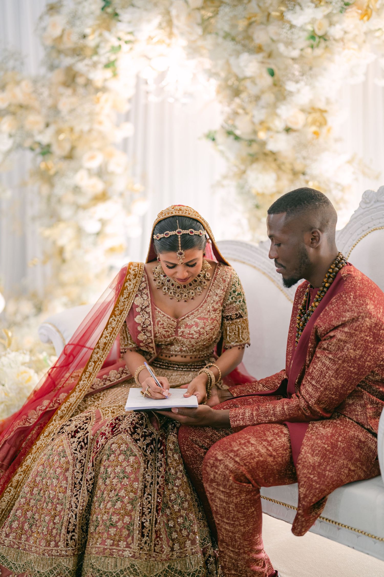 A couple in ornate red and gold South Asian wedding attire share intimate moments on an elegant white sofa.