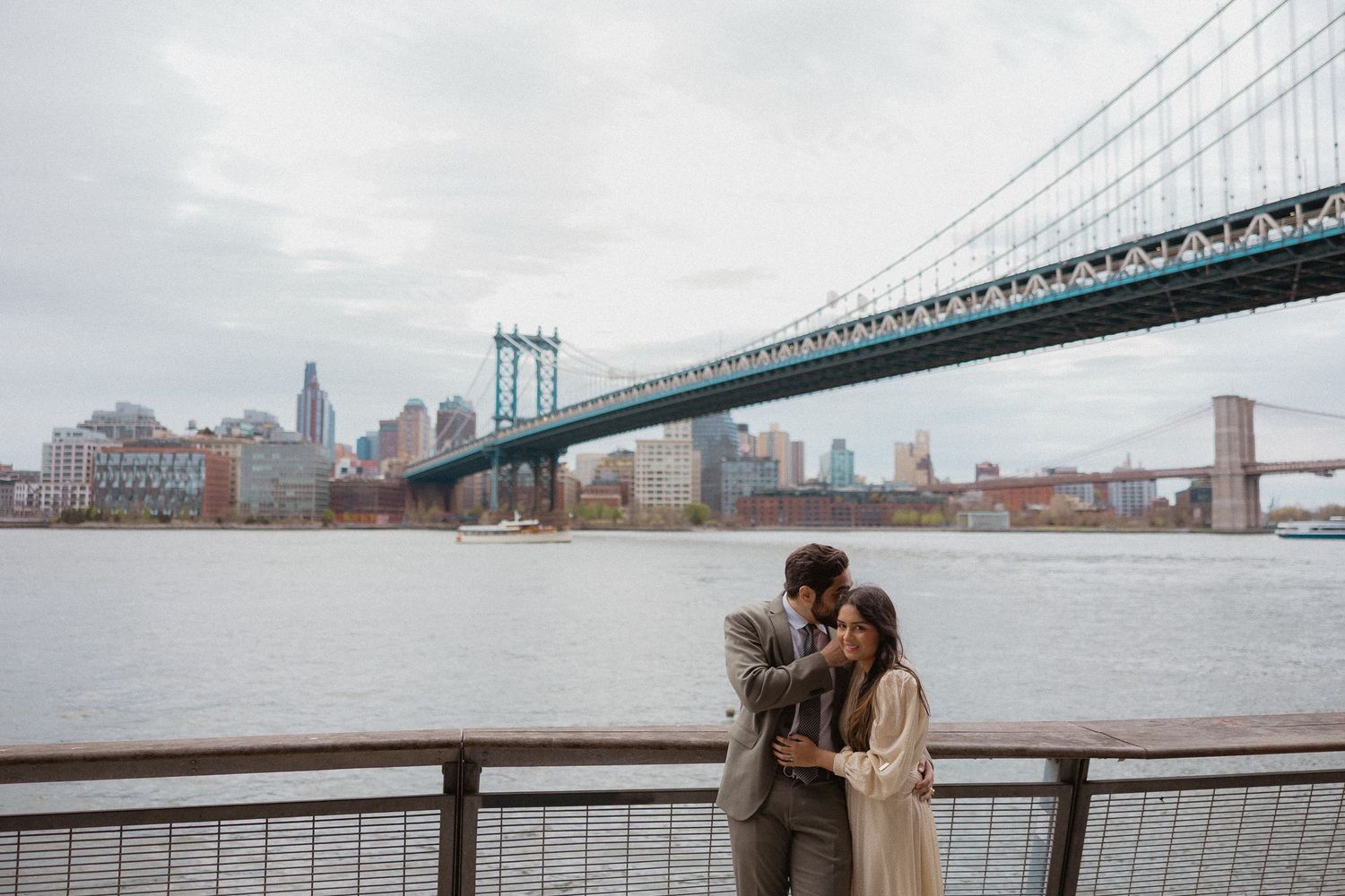 Couple embracing on NYC waterfront with Manhattan Bridge and Brooklyn skyline in background on cloudy day.