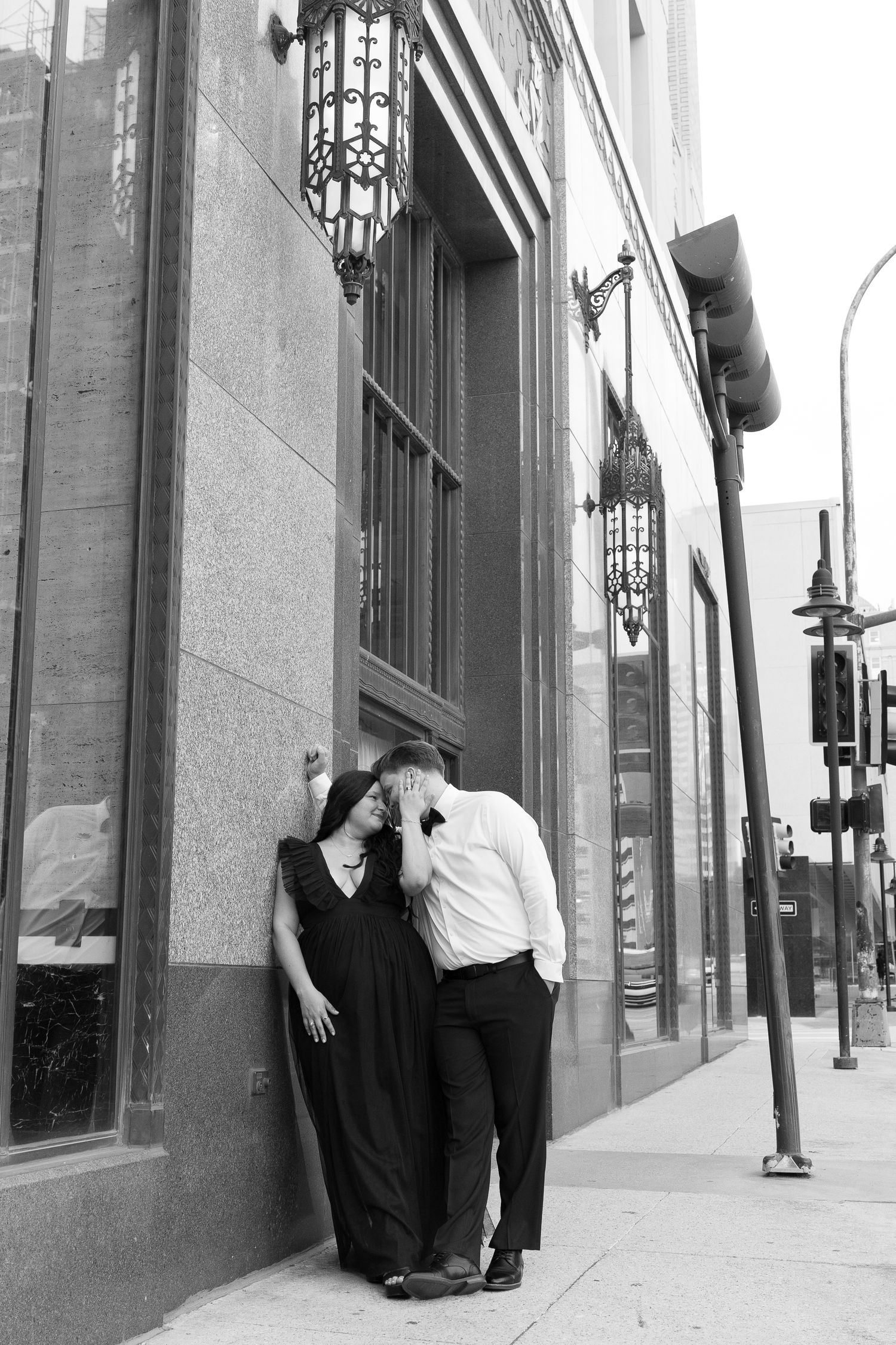 Black and white photo of couple leaning intimately against a stone building facade on a city sidewalk.