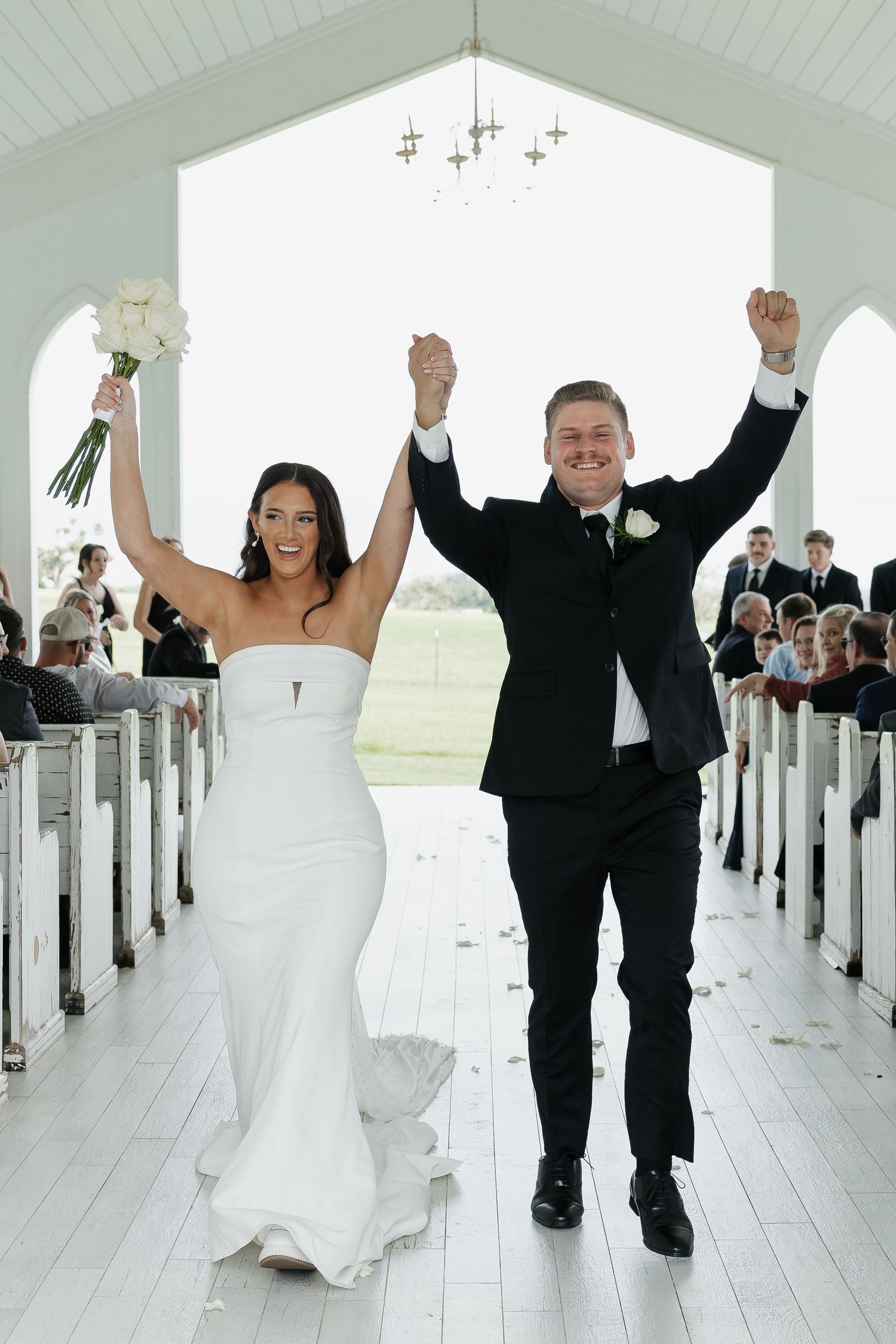 Joyful bride and groom raise hands while walking down aisle of white chapel after wedding ceremony.