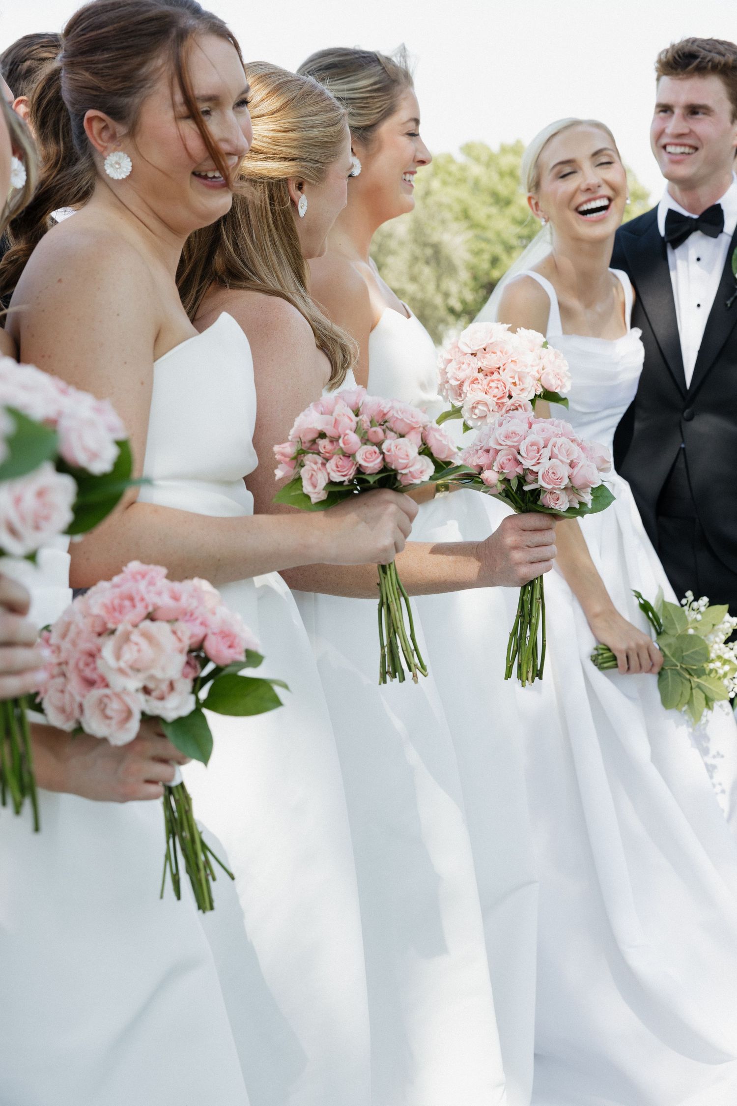 Laughing bridesmaids in white dresses holding pink rose bouquets standing beside bride and groom outdoors.