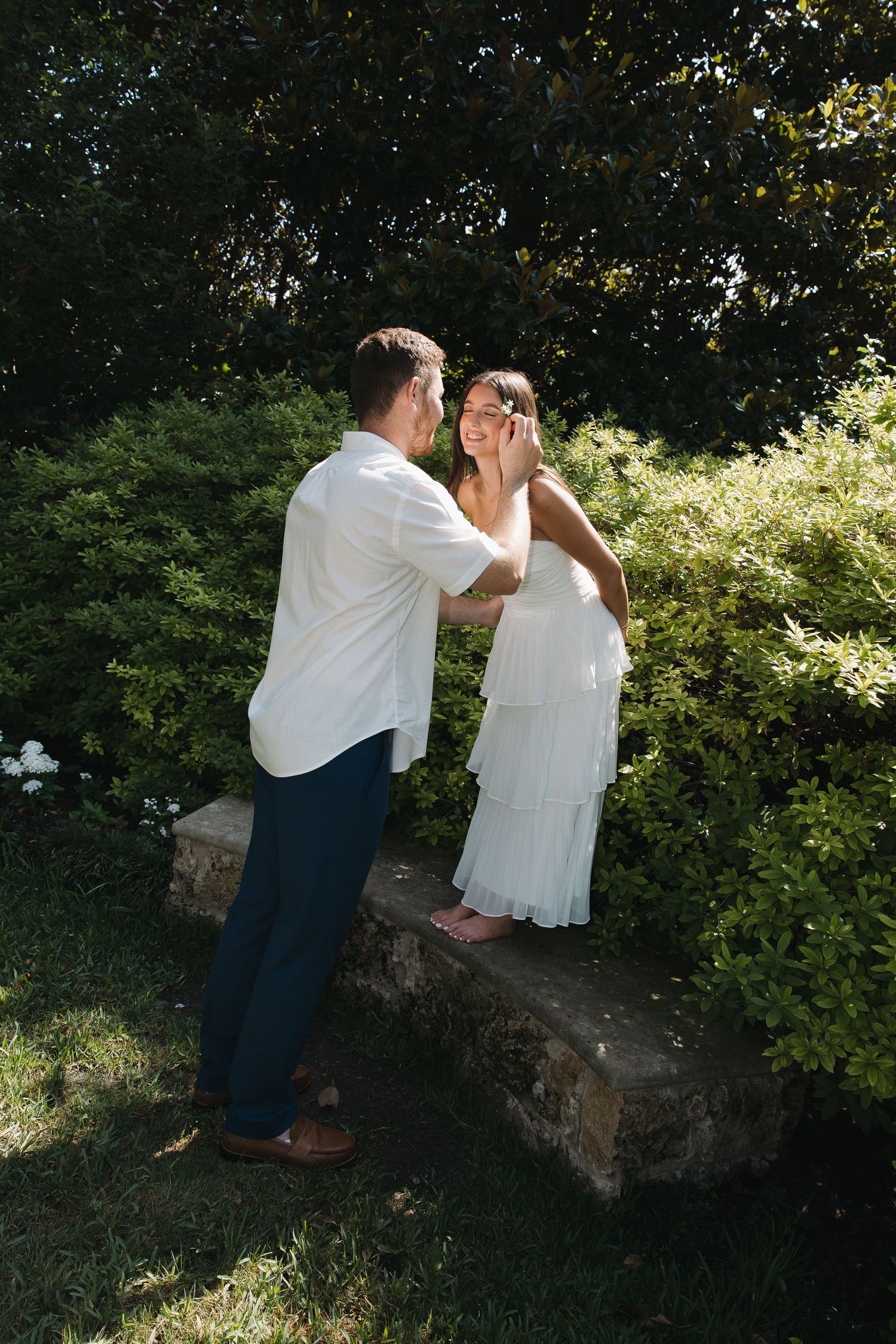 Couple sharing a romantic moment outdoors, bride in white dress and groom in white shirt among lush green garden.