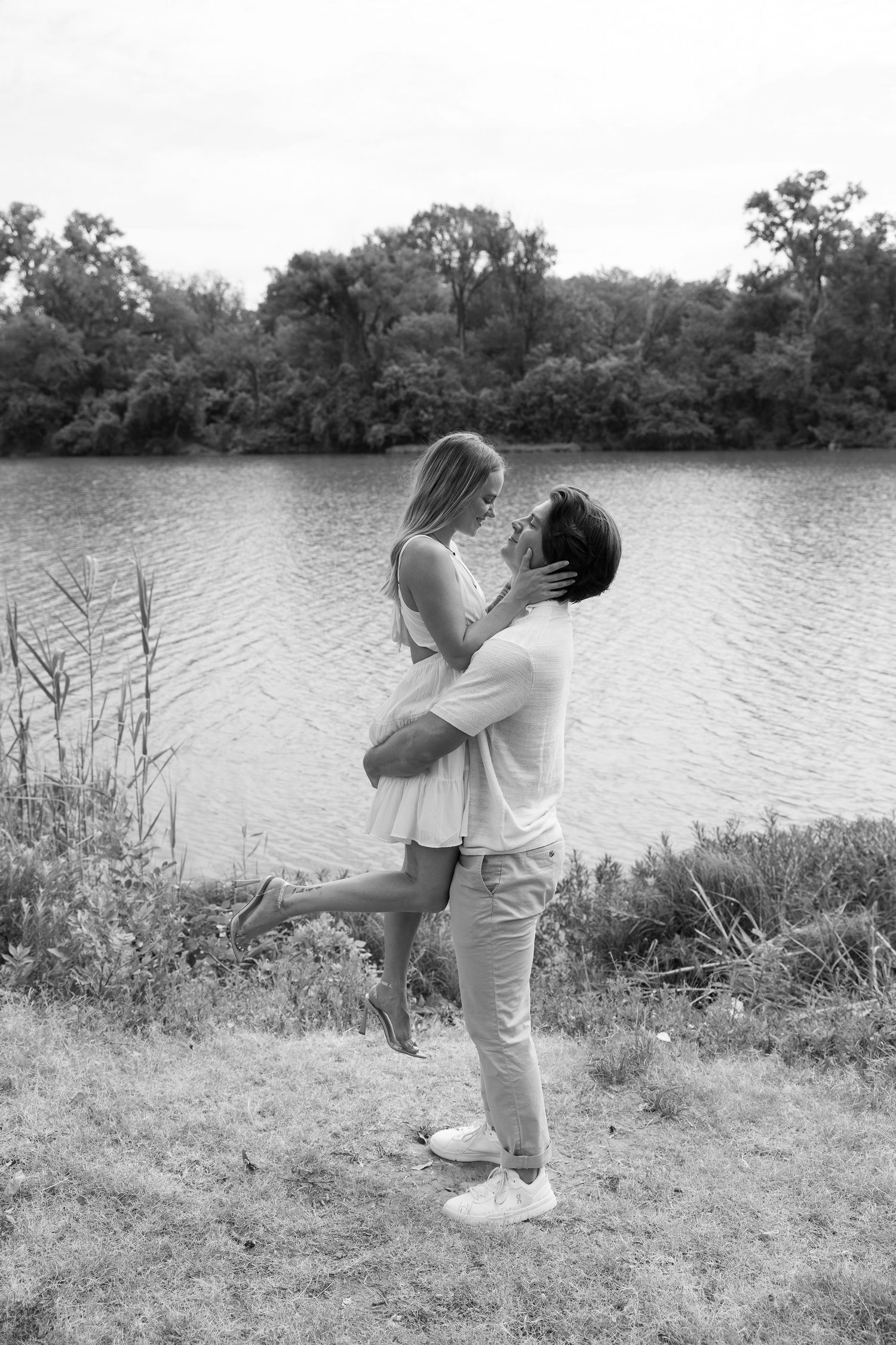Black and white photo of a couple embracing near a lakeside, man lifting woman as they share a romantic moment outdoors.