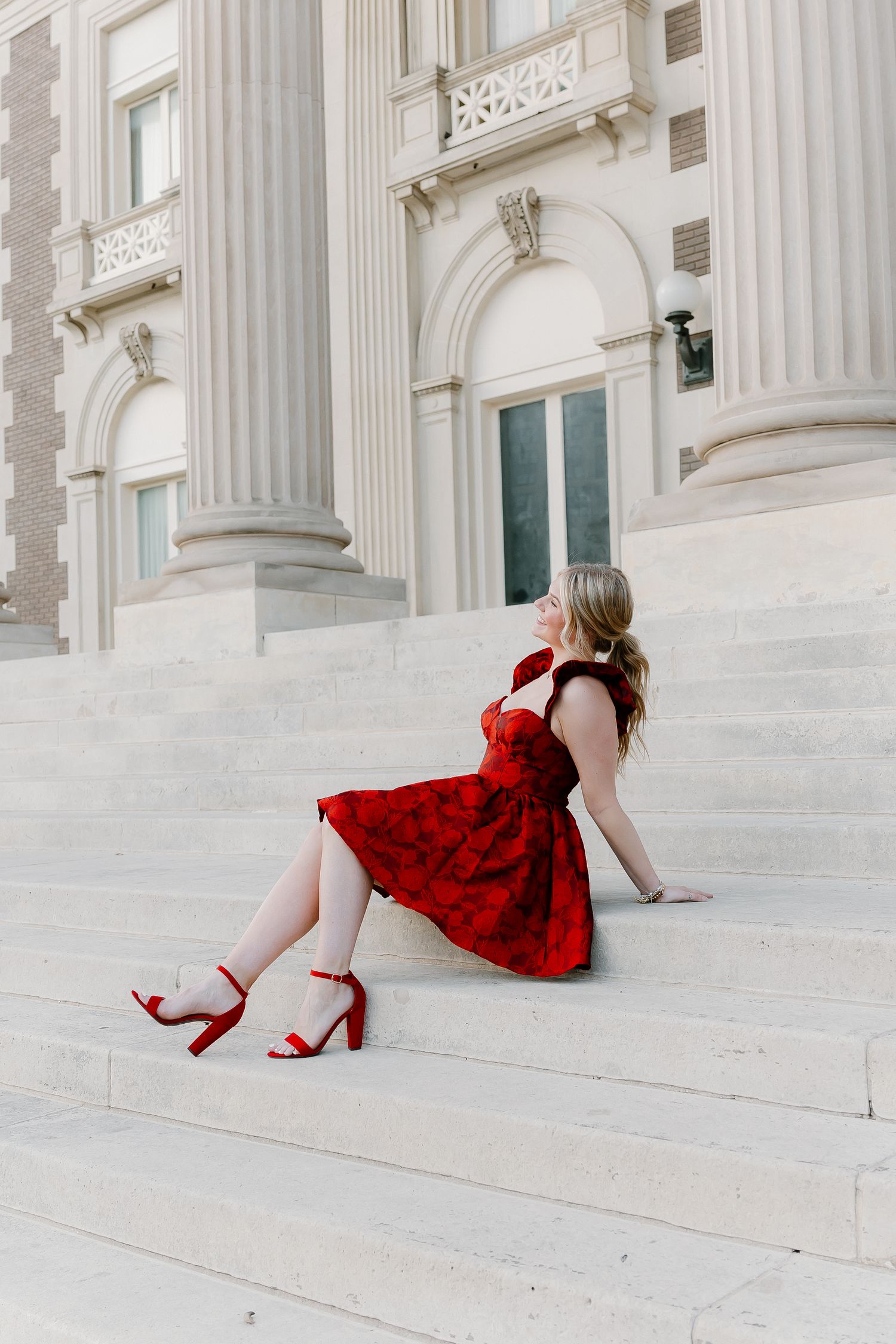 Woman in red floral dress and red block heels sitting gracefully on white marble steps of a classical building.