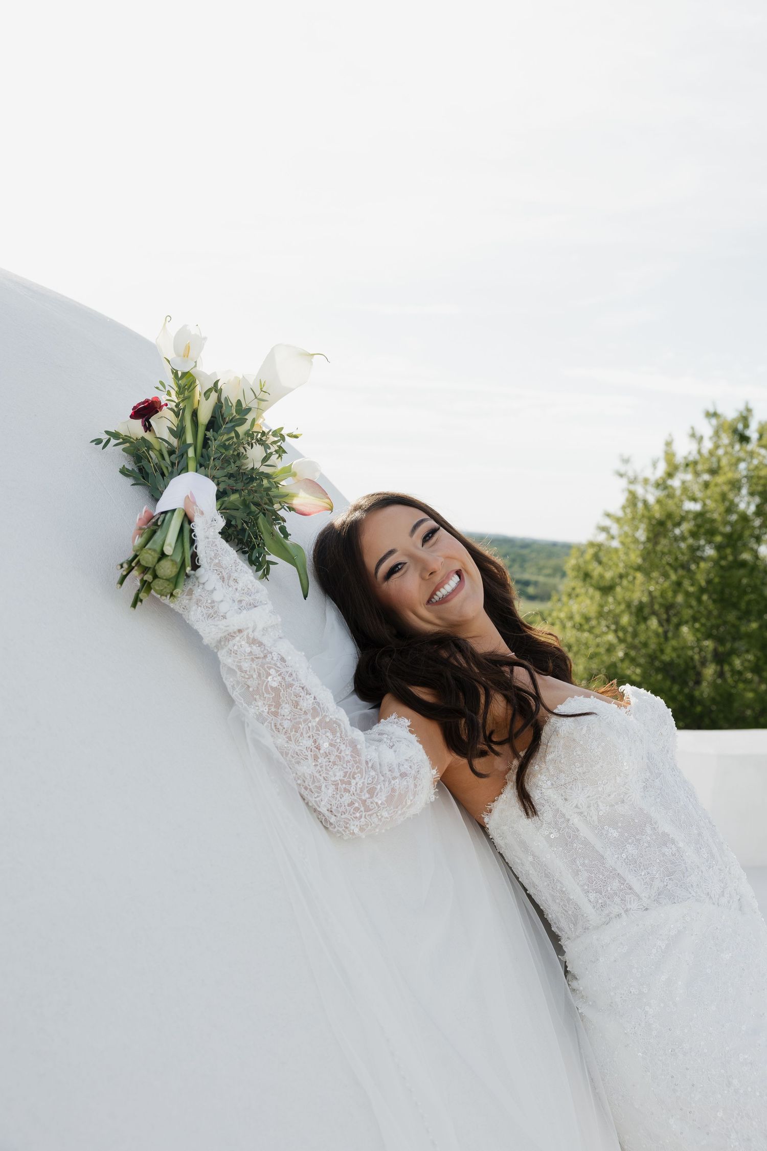 Smiling bride in lace dress holding white floral bouquet leaning against white wall outdoors.