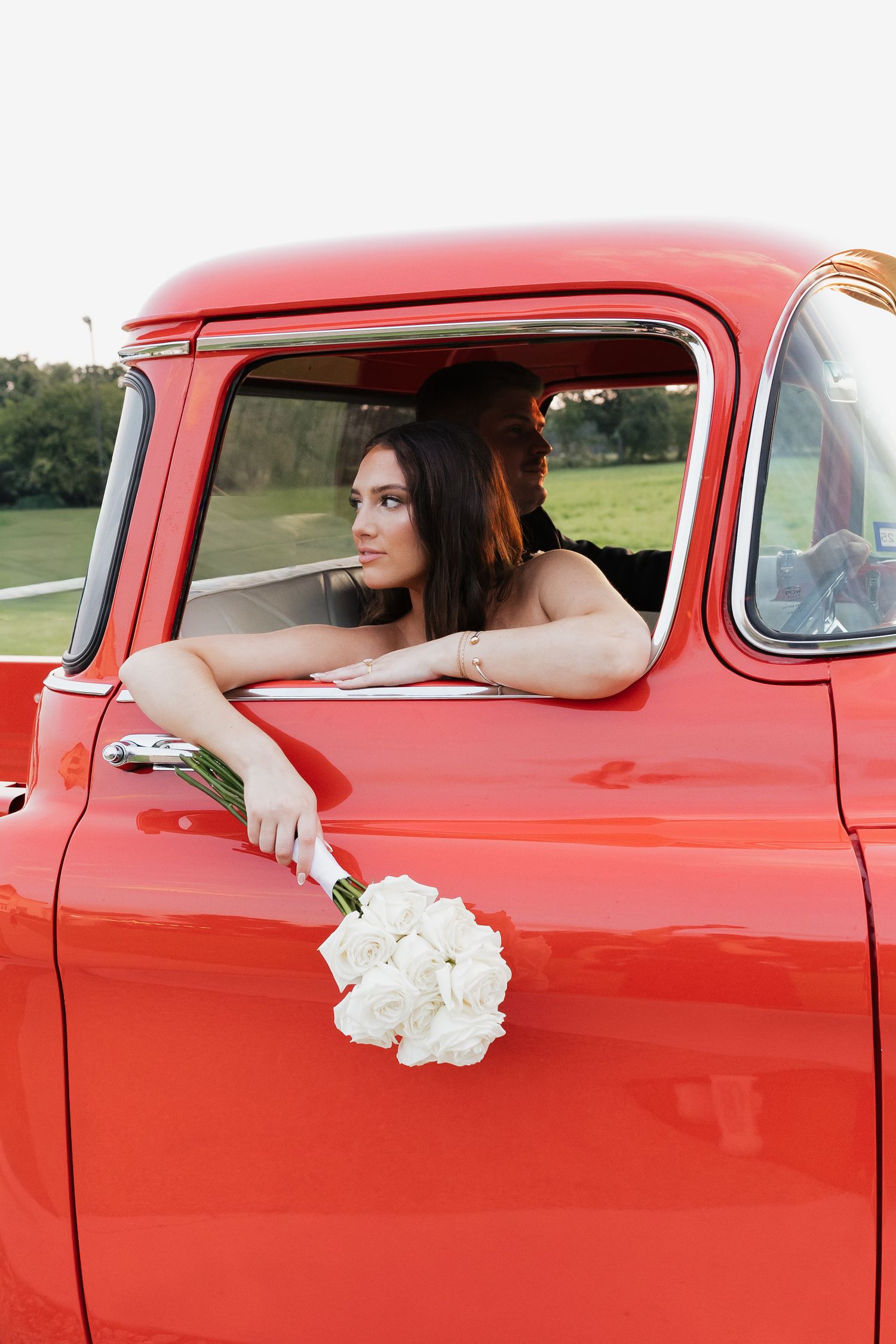 Bride in white wedding dress holds bouquet out window of vintage red pickup truck in outdoor setting.