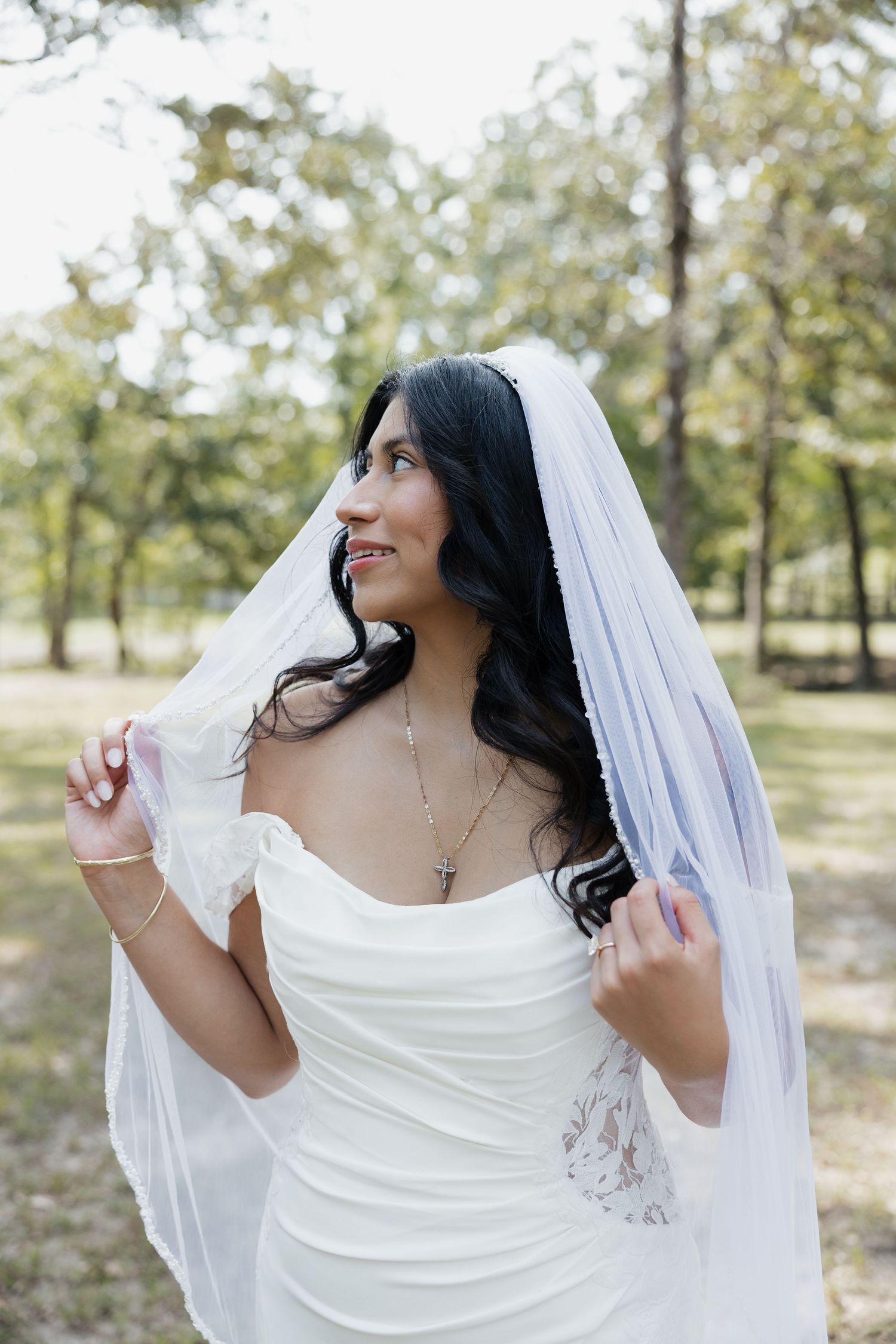 Dark-haired bride in strapless white gown holds veil while smiling in a wooded outdoor setting.