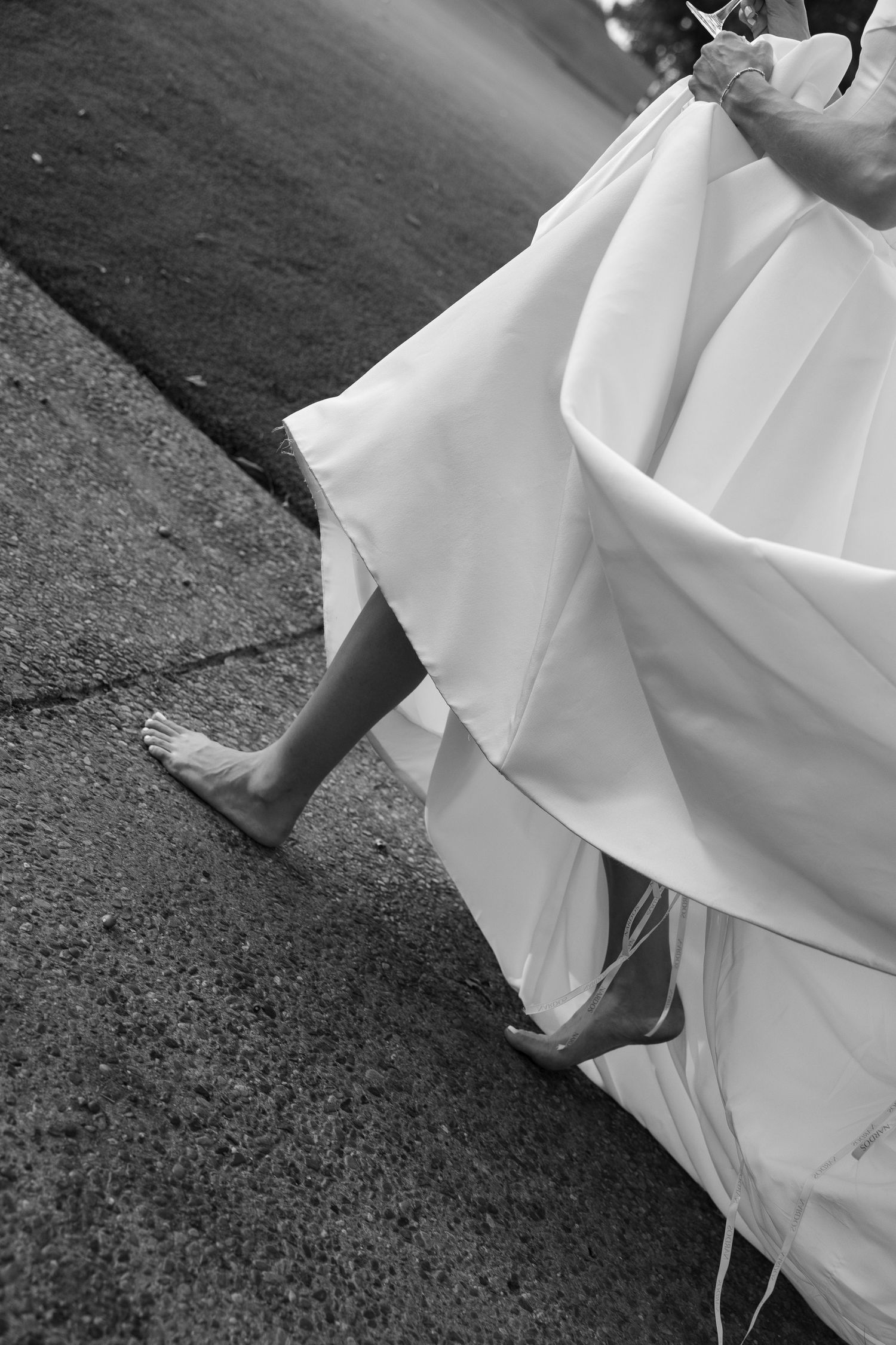 Black and white photo of barefoot bride lifting white wedding gown hem while walking on pavement.