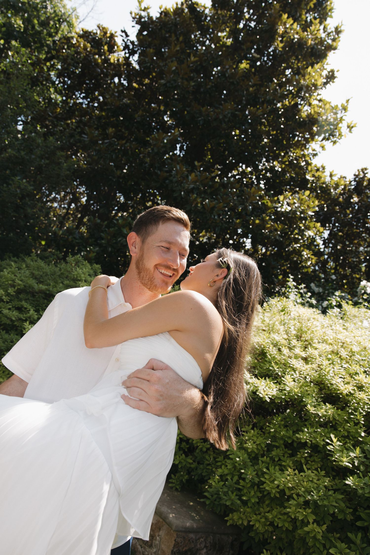 A joyful groom in white lifts his bride outdoors, surrounded by lush green trees on a sunny day.