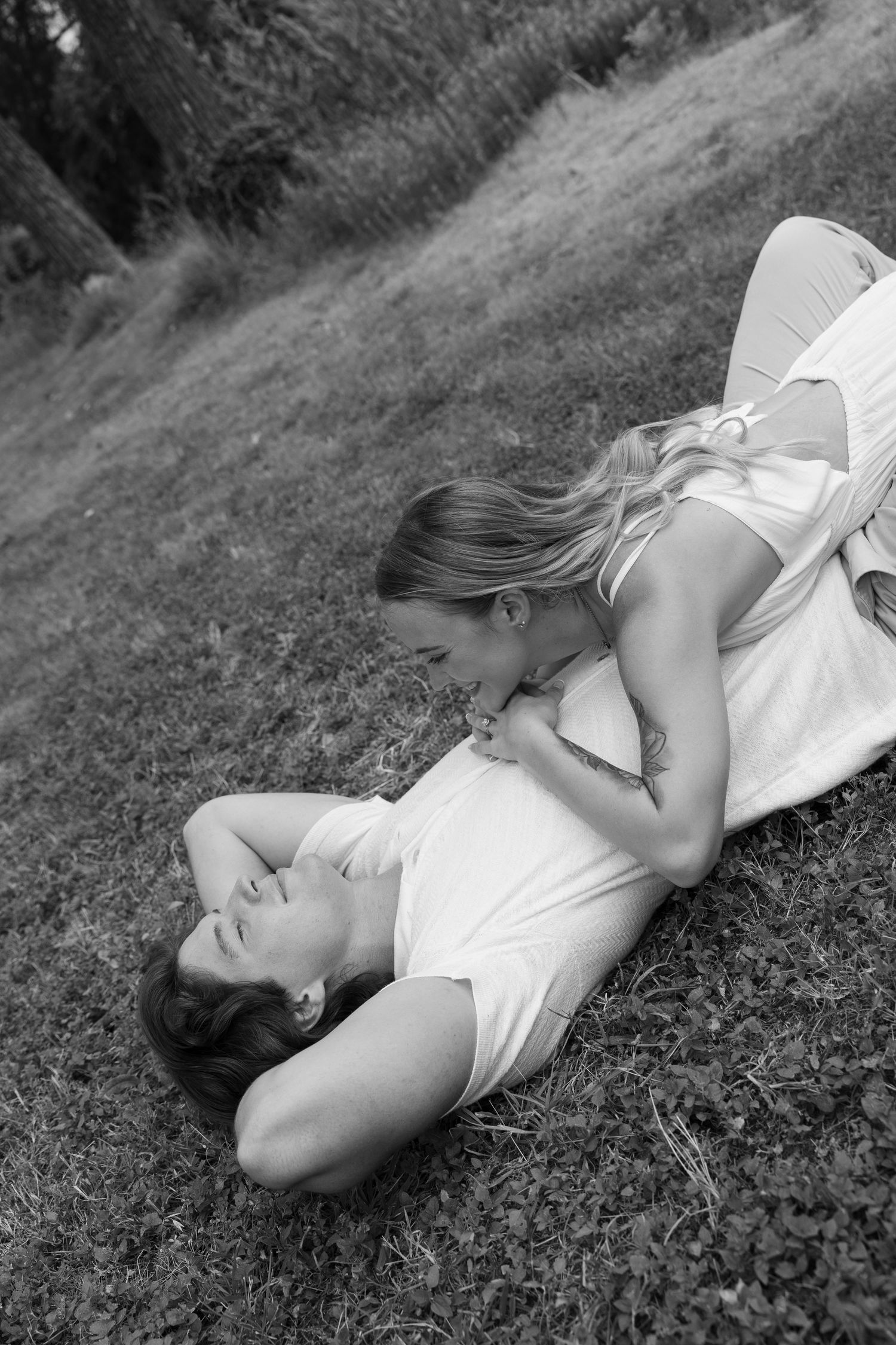 Black and white photo of two women laughing and embracing while lying on grass outdoors.