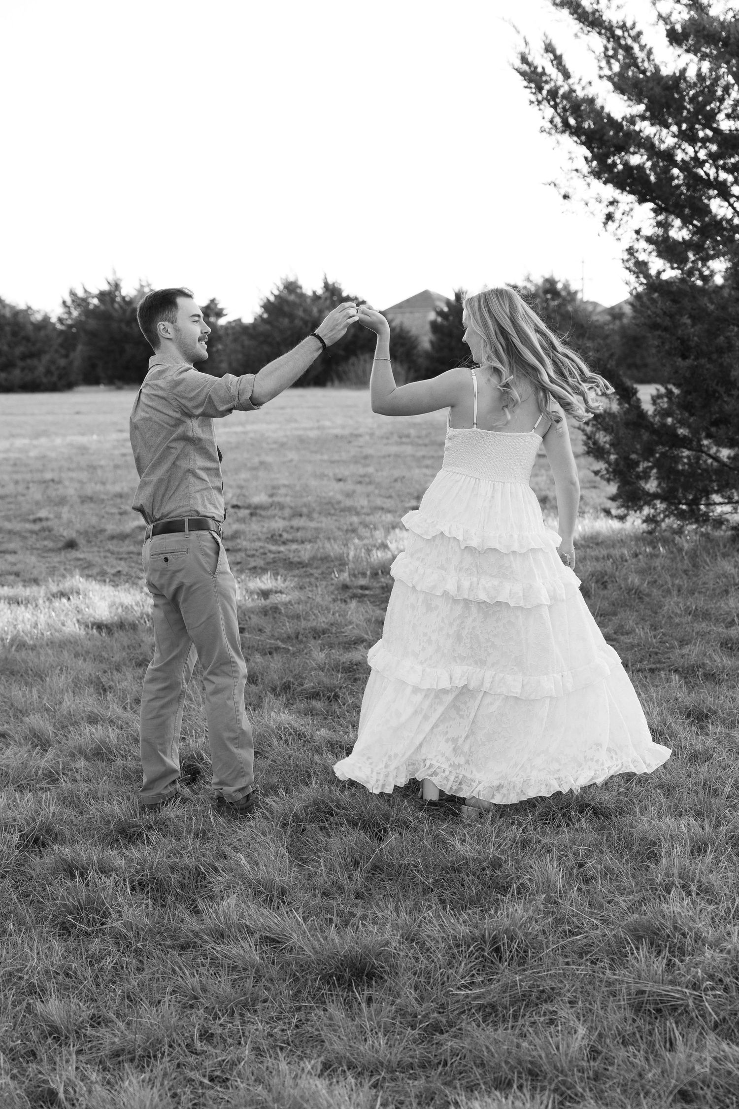 Black and white photo of a couple dancing in a field, bride in ruffled white dress being twirled by groom.