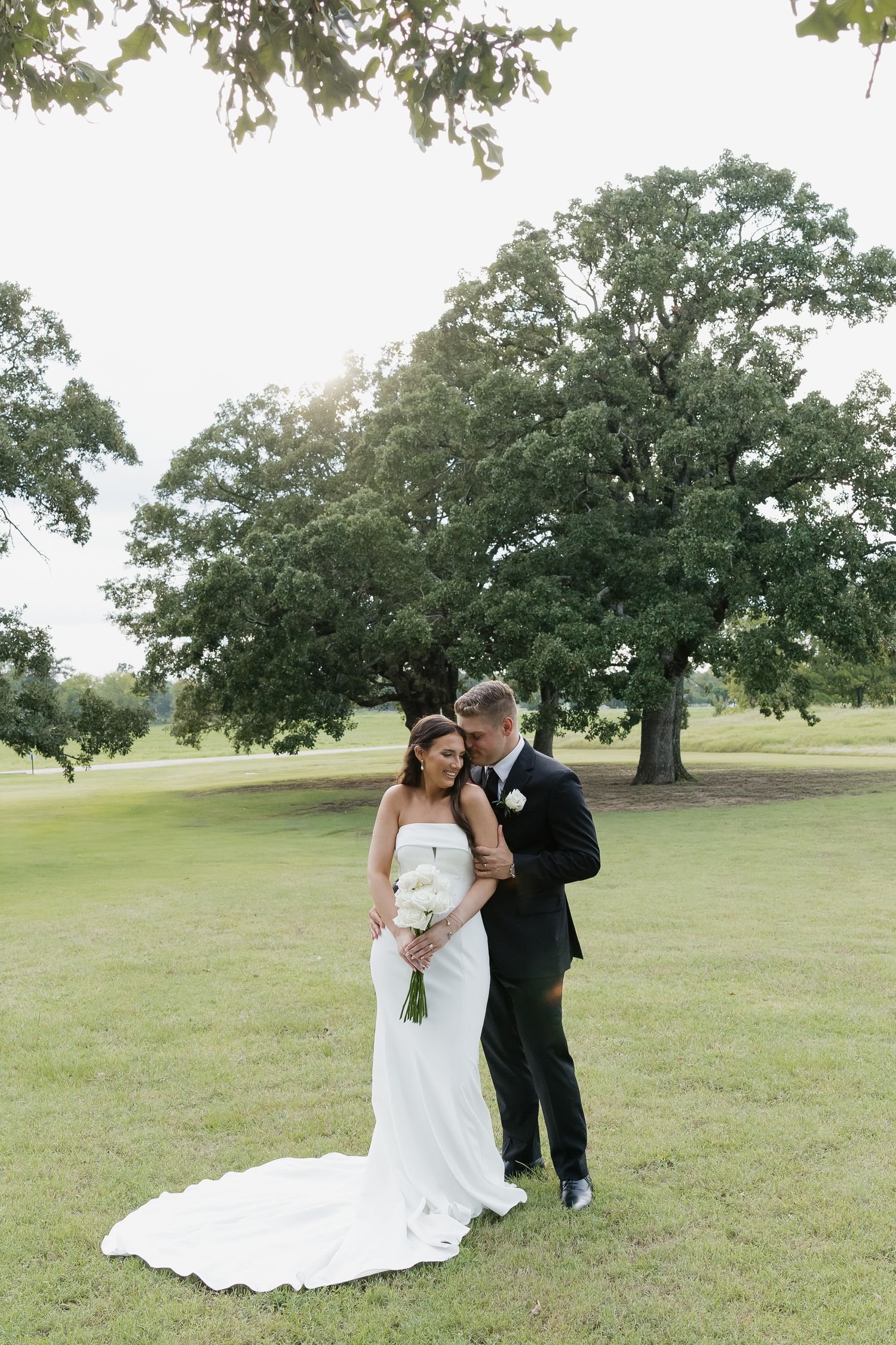 Romantic bride and groom embracing on lush green lawn beneath a large oak tree at golden hour.